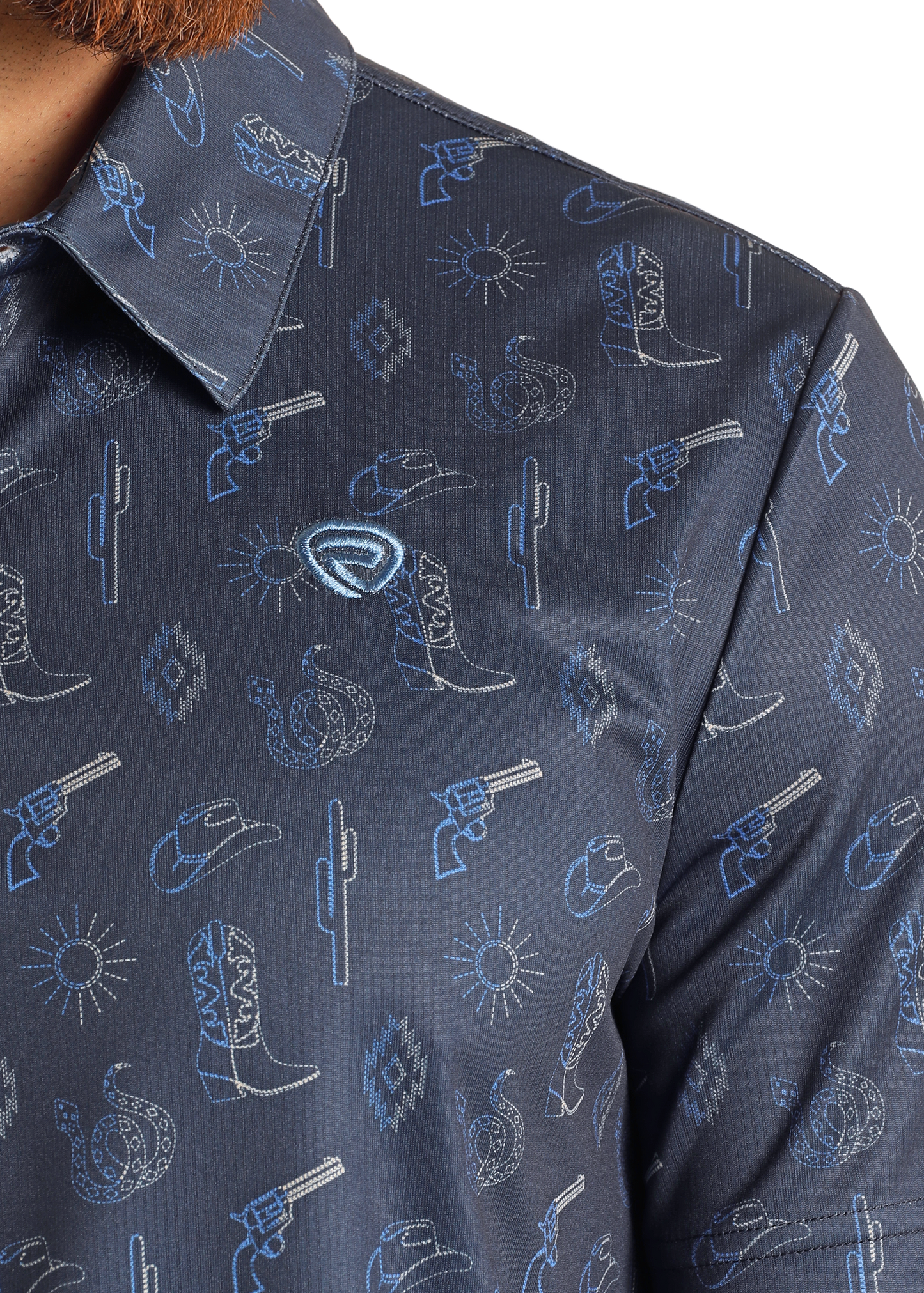 Close-up of the Rock & Roll Denim Cowboy Stitch Printed Polo in navy blue, featuring blue and white outlines of cowboy hats, boots, revolvers, cacti, suns, and a bandana. The collar and part of the wearer's chin are partially visible.
