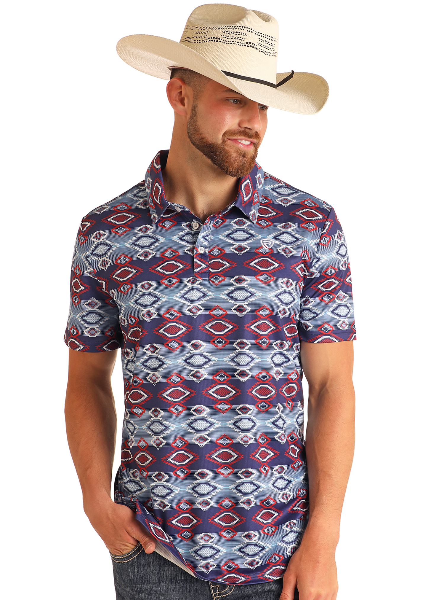 A man wearing a wide-brimmed straw cowboy hat and a Rock & Roll Denim Navy Aztec Print Polo with red, blue, and white designs stands smiling with one hand in his pocket, looking down.