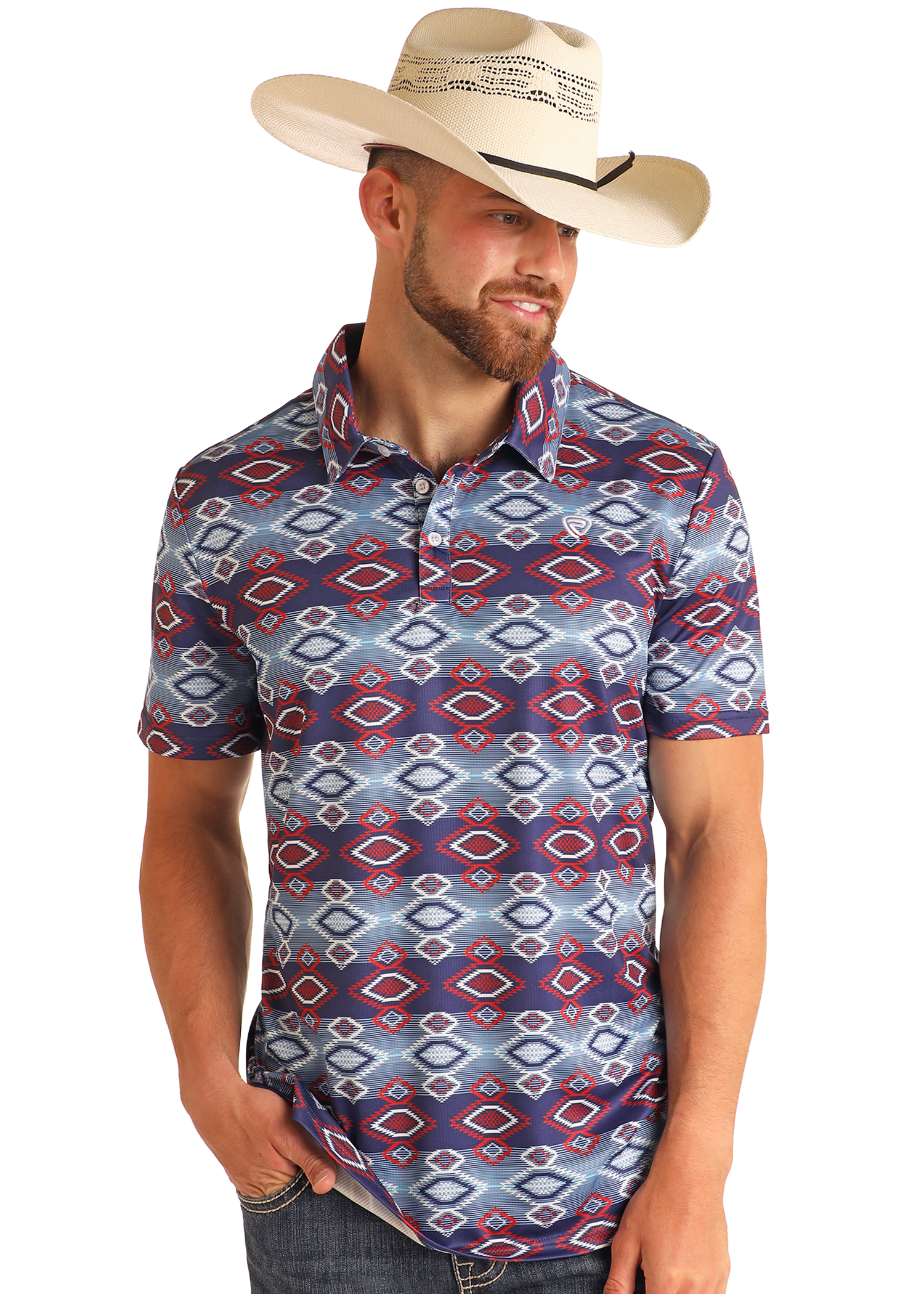 A man wearing a wide-brimmed straw cowboy hat and a Rock & Roll Denim Navy Aztec Print Polo with red, blue, and white designs stands smiling with one hand in his pocket, looking down.