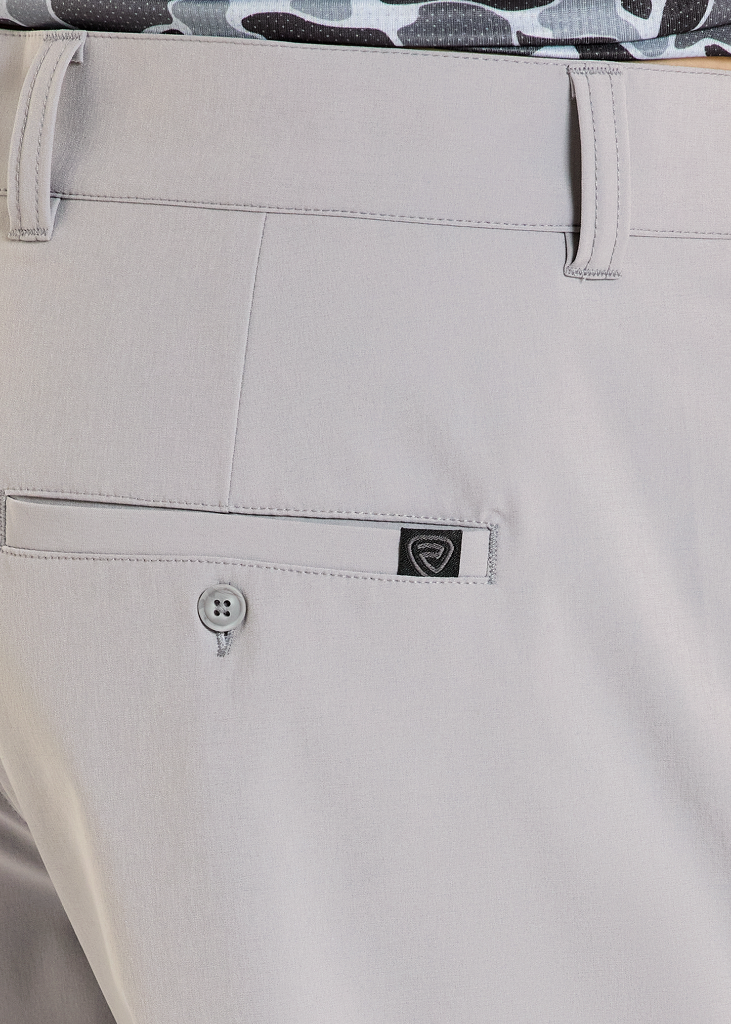Close-up of beige pants with a brand logo on the pocket, featuring a camouflage shirt in the background.