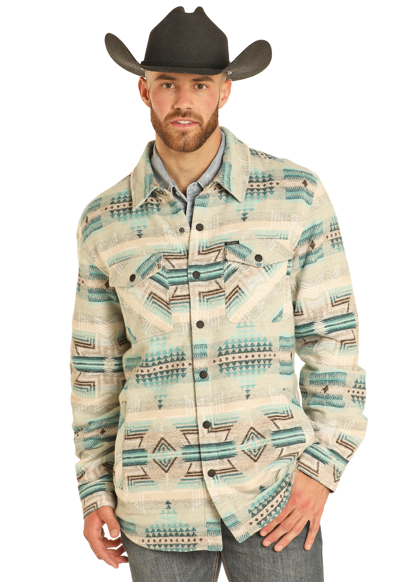 A man wearing a black cowboy hat, light blue shirt, gray jeans, and the Rock & Roll Denim Aztec Print Jacquard Shirt Jacket featuring cream, teal, beige, and black geometric designs stands facing forward with one hand in his pocket.