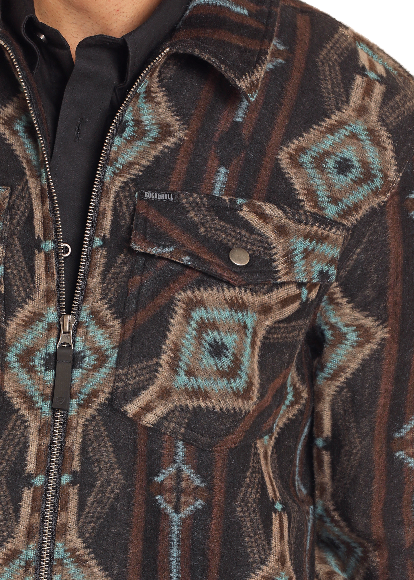 A close-up of a person wearing the Rock & Roll Denim Aztec Print Brushed Jacquard Zip Up Jacket, featuring brown, black, and teal geometric designs, a zippered front, snap-button chest pocket, and layered over a black collared shirt.