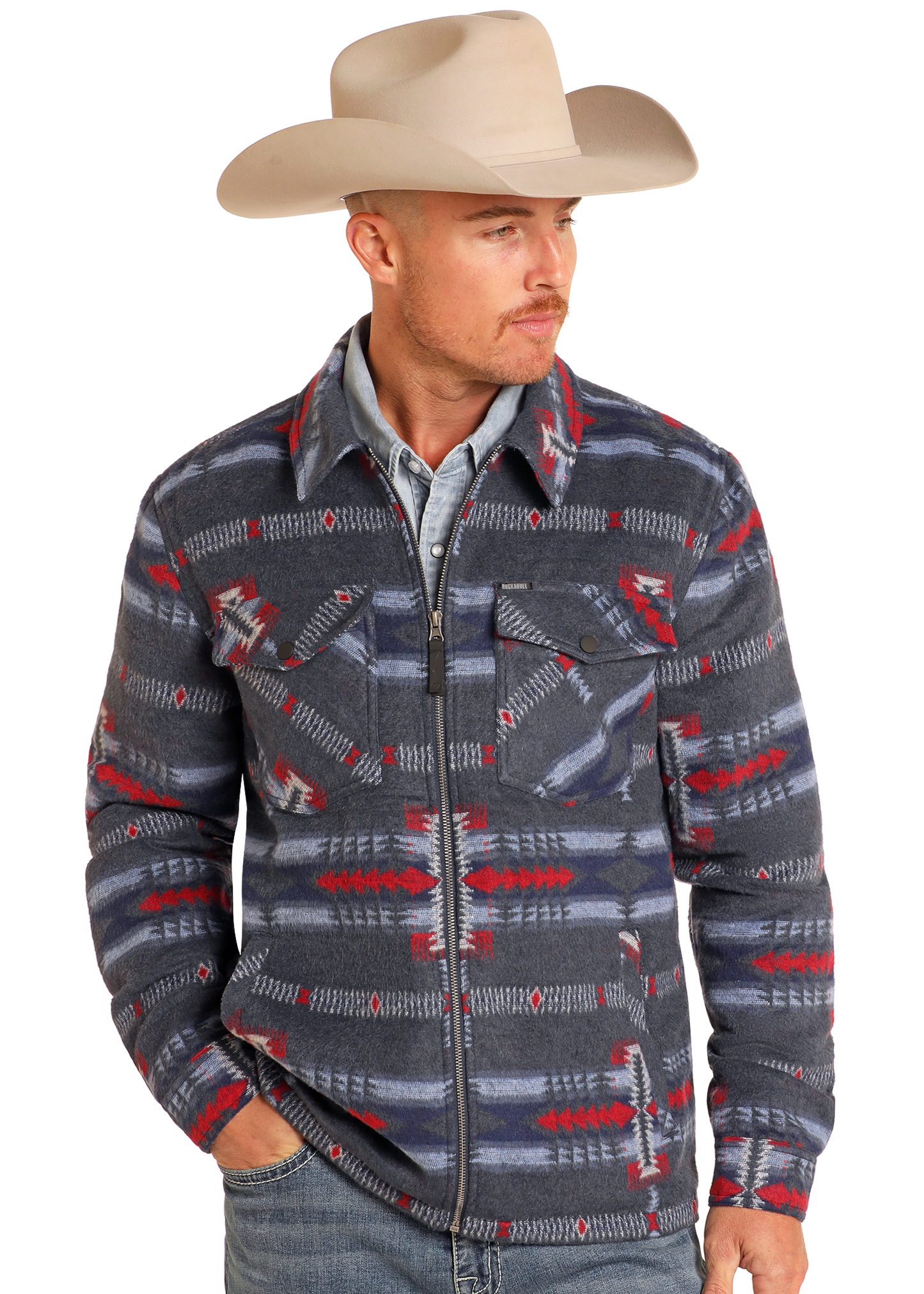 A man wearing a light tan cowboy hat, Rock & Roll Denim Blue Aztec Brushed Jacquard Zip Up Jacket, a light denim shirt, and blue jeans stands with one hand in his pocket, looking to the side.