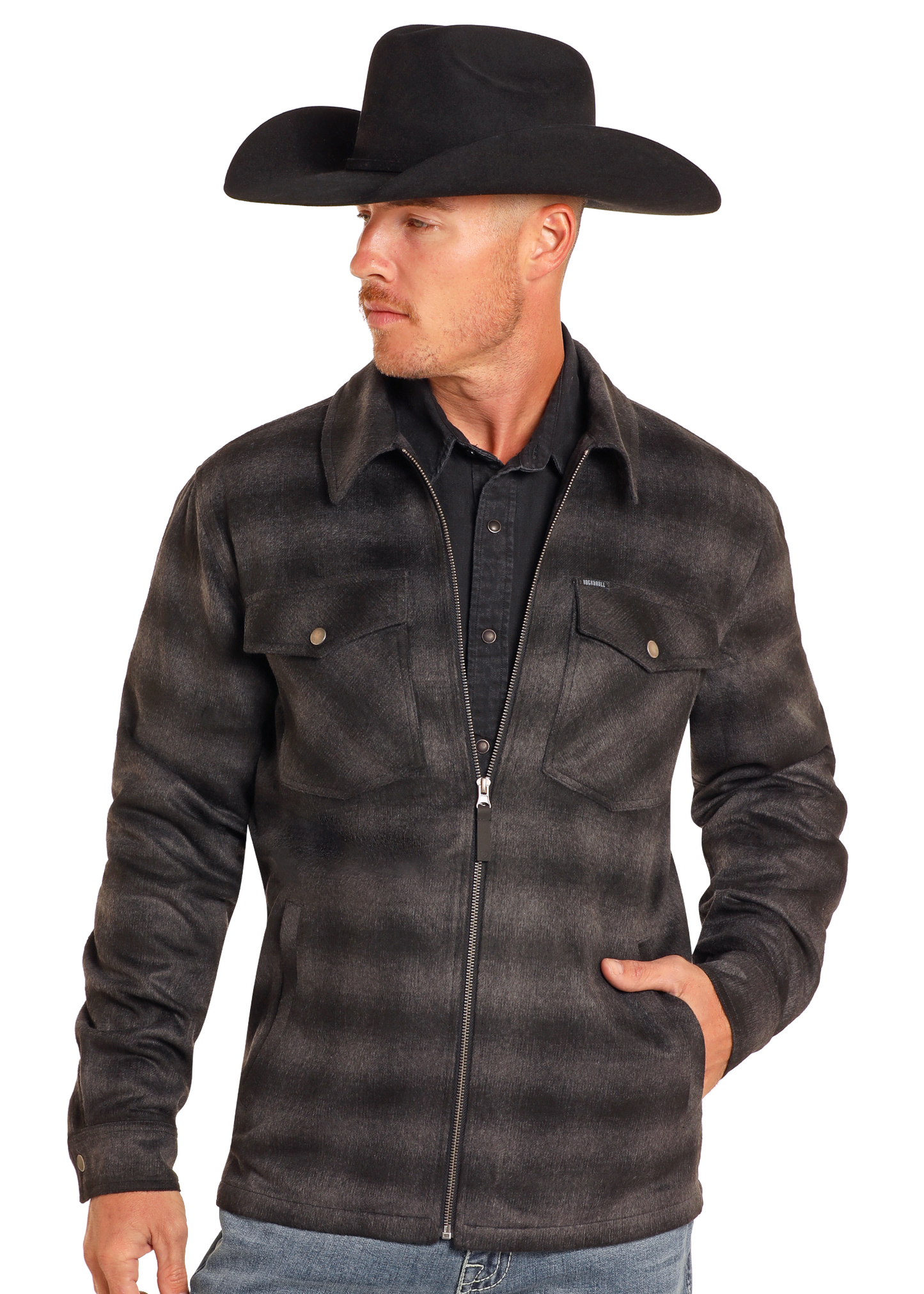 A man wearing a black cowboy hat, black shirt, blue jeans, and the Rock & Roll Denim Brushed Ombre Plaid Zip Up Jacket with buttoned chest pockets stands with one hand in his jacket pocket, looking to his left.