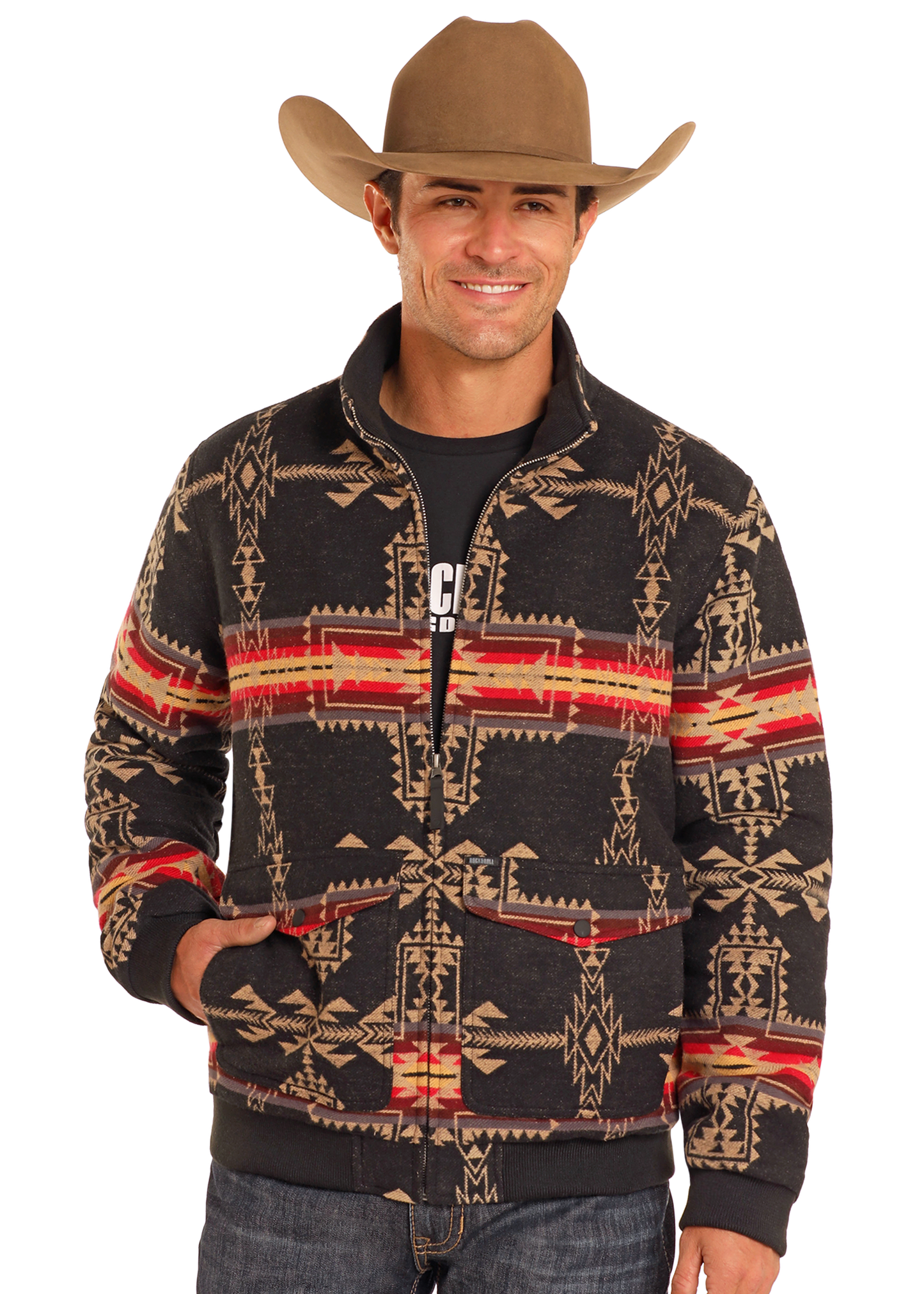 A smiling man, wearing the Rock & Roll Denim Large Aztec Jacquard Bomber Jacket with red and tan geometric designs over a black shirt, blue jeans, and a brown cowboy hat, poses with one hand in his pocket.