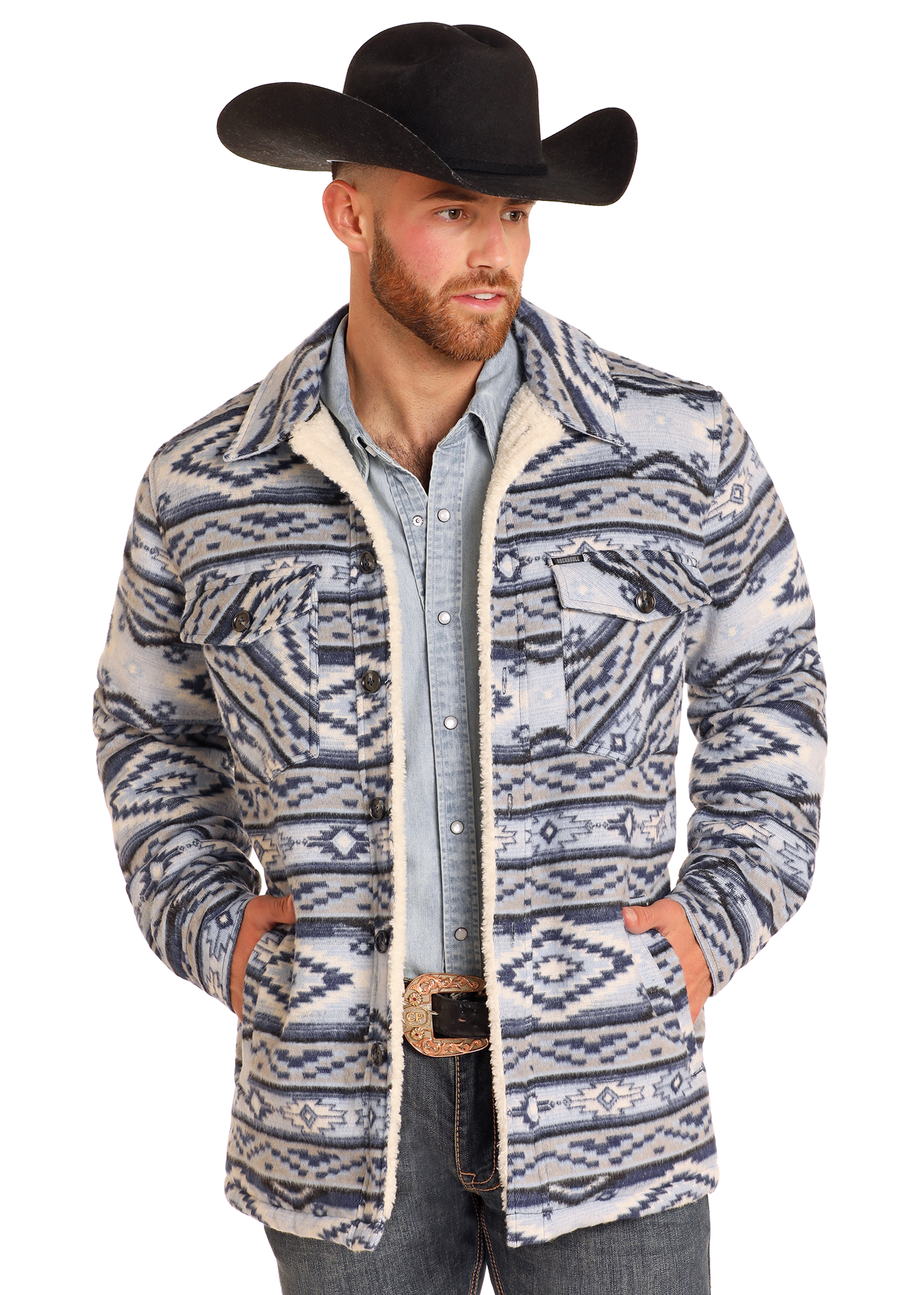 A man wearing a black cowboy hat, Rock & Roll Denim Aztec Brushed Jacquard Shirt Jacket, denim shirt, jeans, and decorative belt buckle stands with his hands in his jacket pockets.