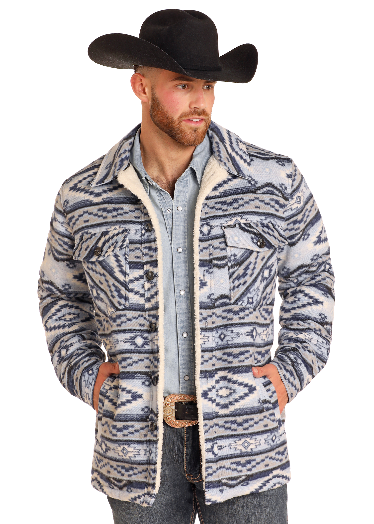 A man wearing a black cowboy hat, Rock & Roll Denim Aztec Brushed Jacquard Shirt Jacket, denim shirt, jeans, and decorative belt buckle stands with his hands in his jacket pockets.