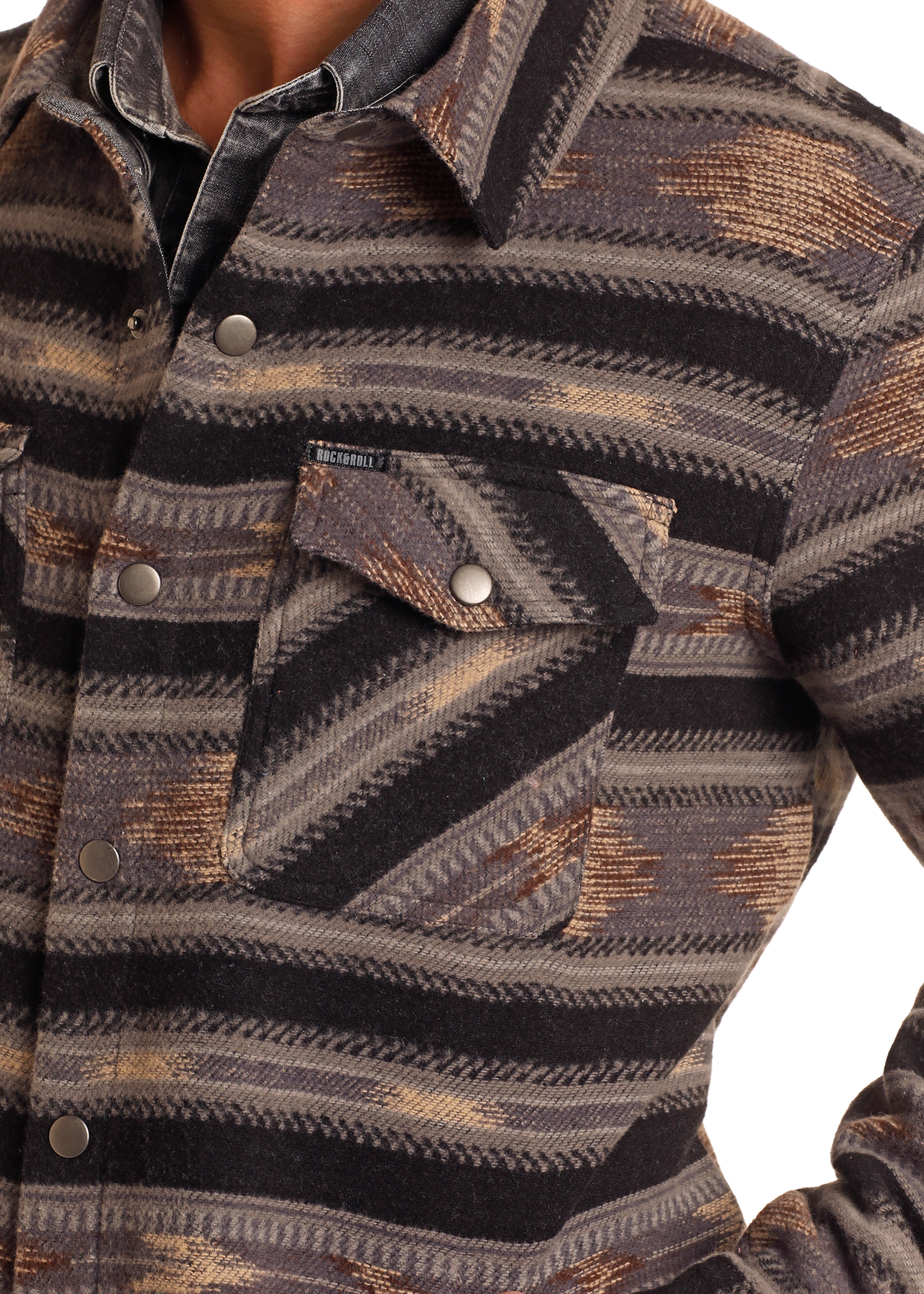 A man wears the Rock & Roll Denim Aztec Striped Jacquard Shirt Jacket, featuring a southwestern geometric pattern in brown, gray, and black. Made from 100% cotton with a chest flap pocket and snap button closure.