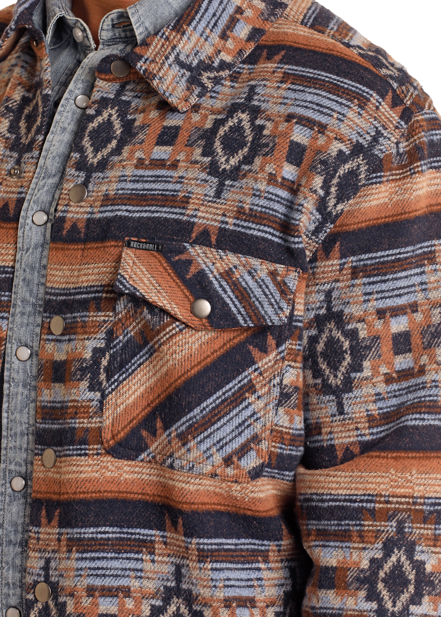 A close-up of a person wearing the Rock & Roll Denim Navy Aztec Striped Jacquard Shirt Jacket over a light denim shirt. Crafted from 100% cotton, it has a front pocket with button flap and metal snap buttons down the front.