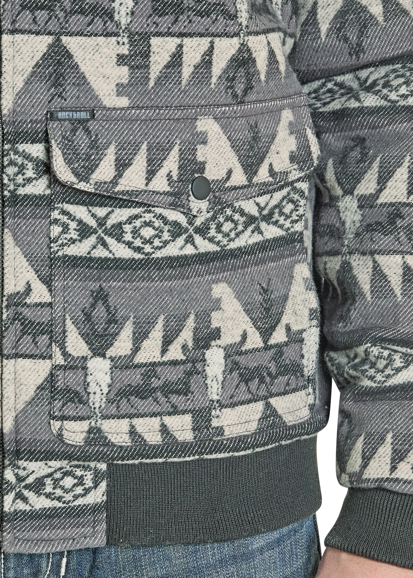 A close-up of the Rock & Roll Denim Jacquard Aztec Horse Bomber Jacket, featuring a gray, black, and cream southwestern geometric pattern, large flap pocket, cotton jacquard fabric, and shown with part of an arm and blue jeans.