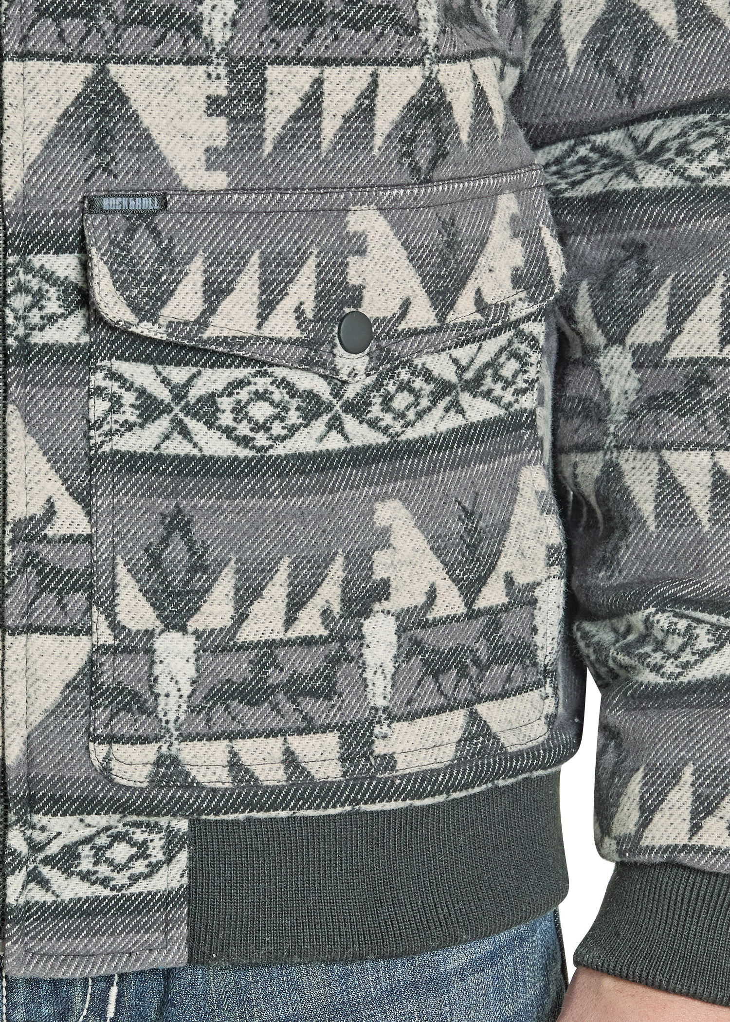 A close-up of the Rock & Roll Denim Jacquard Aztec Horse Bomber Jacket, featuring a gray, black, and cream southwestern geometric pattern, large flap pocket, cotton jacquard fabric, and shown with part of an arm and blue jeans.