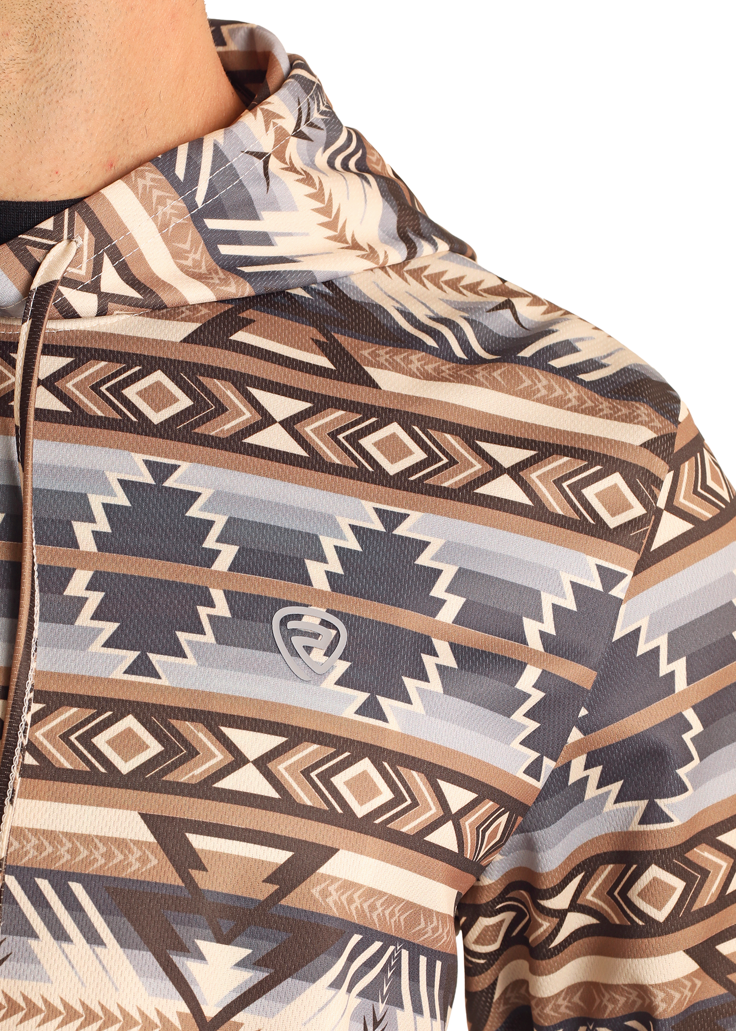 A close-up of someone wearing the Rock & Roll Denim Tan Aztec Performance Hoodie, featuring a geometric southwestern print in brown, tan, and gray with a small gray shield-shaped logo on the chest.