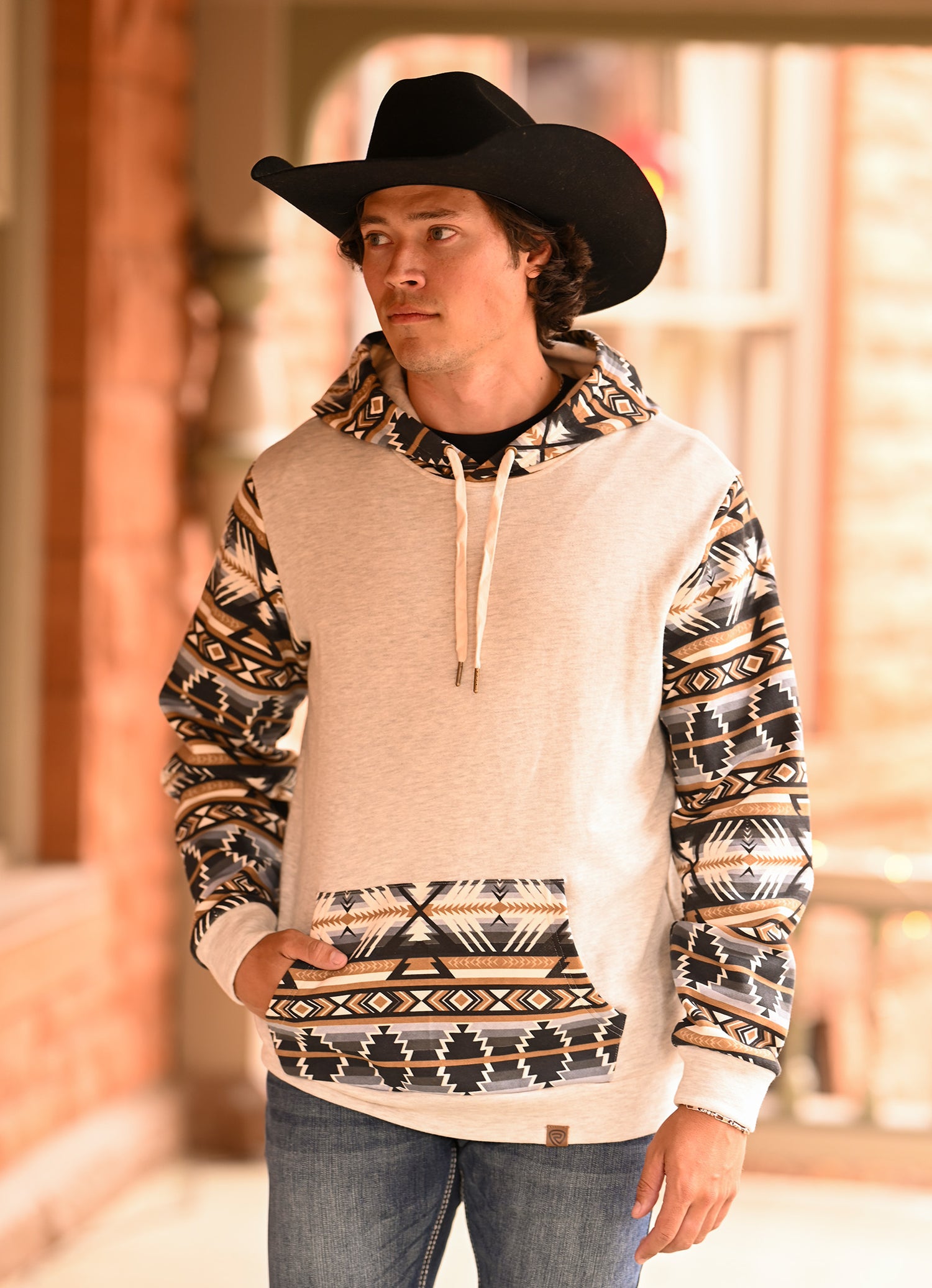 A man in a black cowboy hat, blue jeans, and a Rock & Roll Denim Aztec Accented Hoodie with patterned sleeves and pocket stands outdoors in a rustic setting, looking to the side with one hand in his pocket.