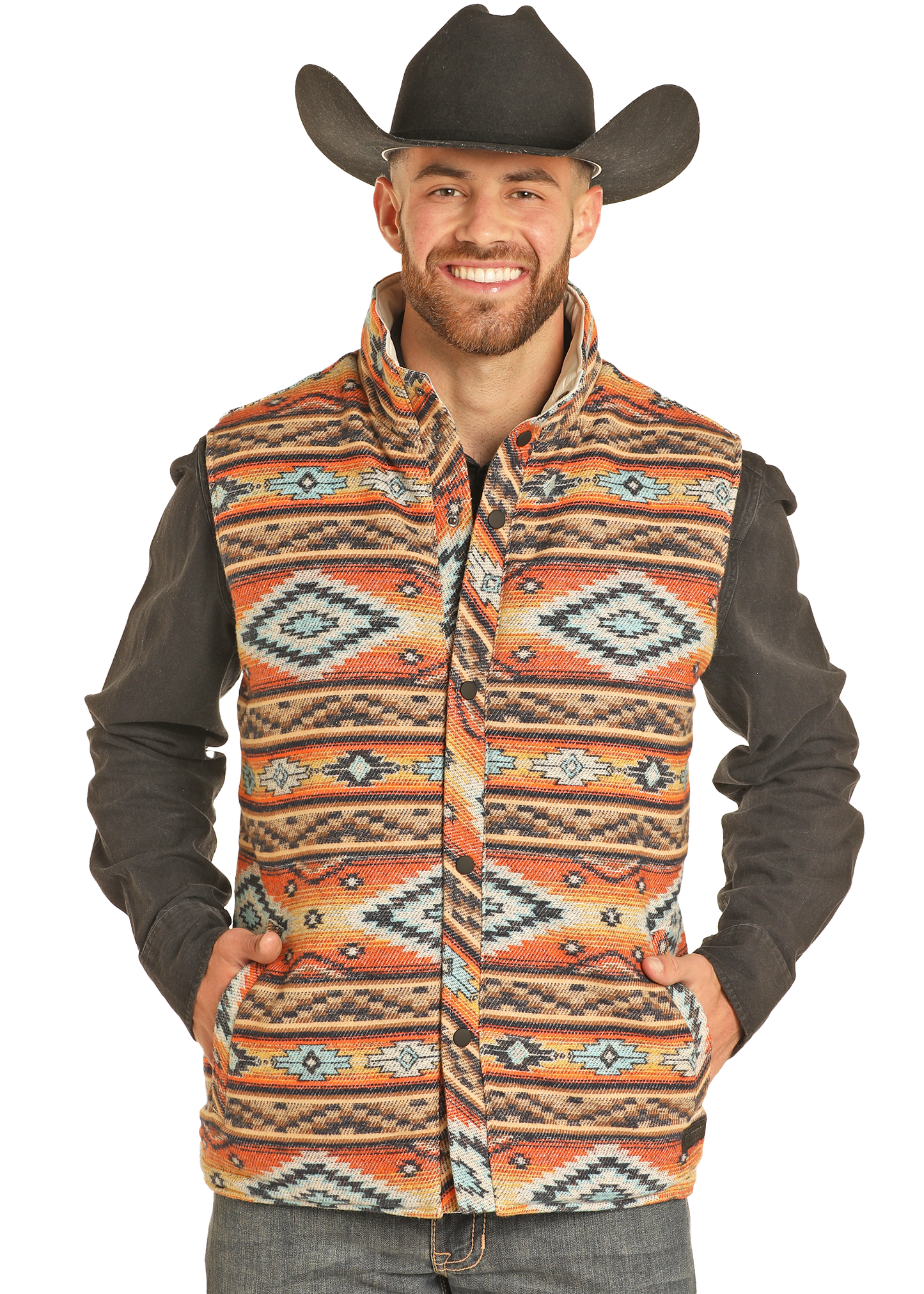 A man smiles as he poses in jeans, a dark shirt, and the Rock & Roll Denim Reversible Aztec Jacquard Vest—featuring bold orange, blue, and beige patterns—topped with a black cowboy hat for standout style.