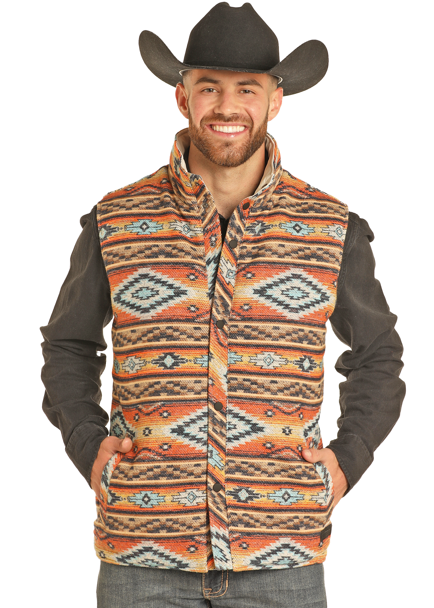 A man smiles as he poses in jeans, a dark shirt, and the Rock & Roll Denim Reversible Aztec Jacquard Vest—featuring bold orange, blue, and beige patterns—topped with a black cowboy hat for standout style.