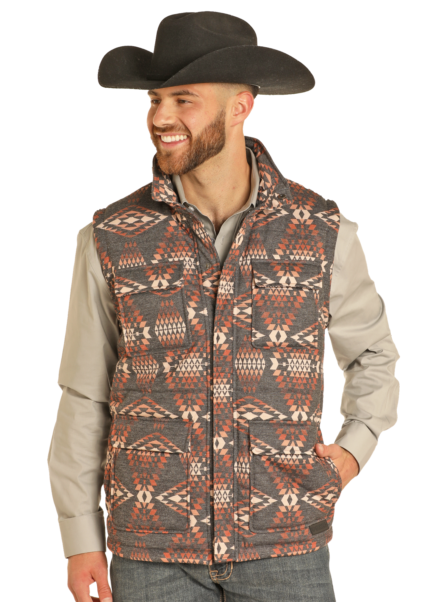 A smiling man in a black cowboy hat, light gray shirt, and Rock & Roll Denim Aztec Puffer Vest with a red and white geometric pattern stands with one hand in his pocket against a plain background.