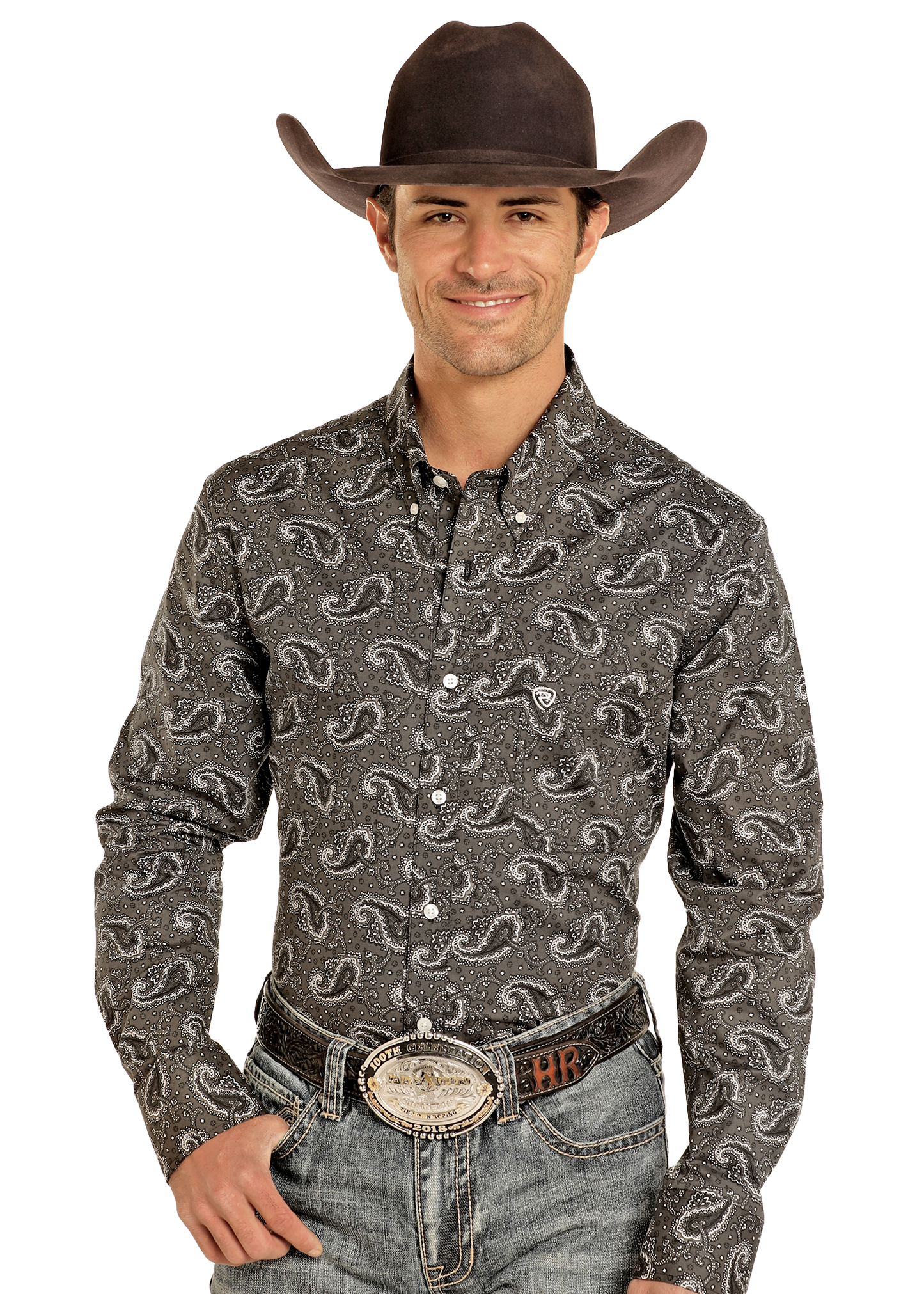 A man wearing a black cowboy hat, Rock & Roll Denim Classic Fit Paisley Print Long Sleeve Button Down Shirt, and blue jeans with a large belt buckle smiles with one hand in his pocket against a plain light background.