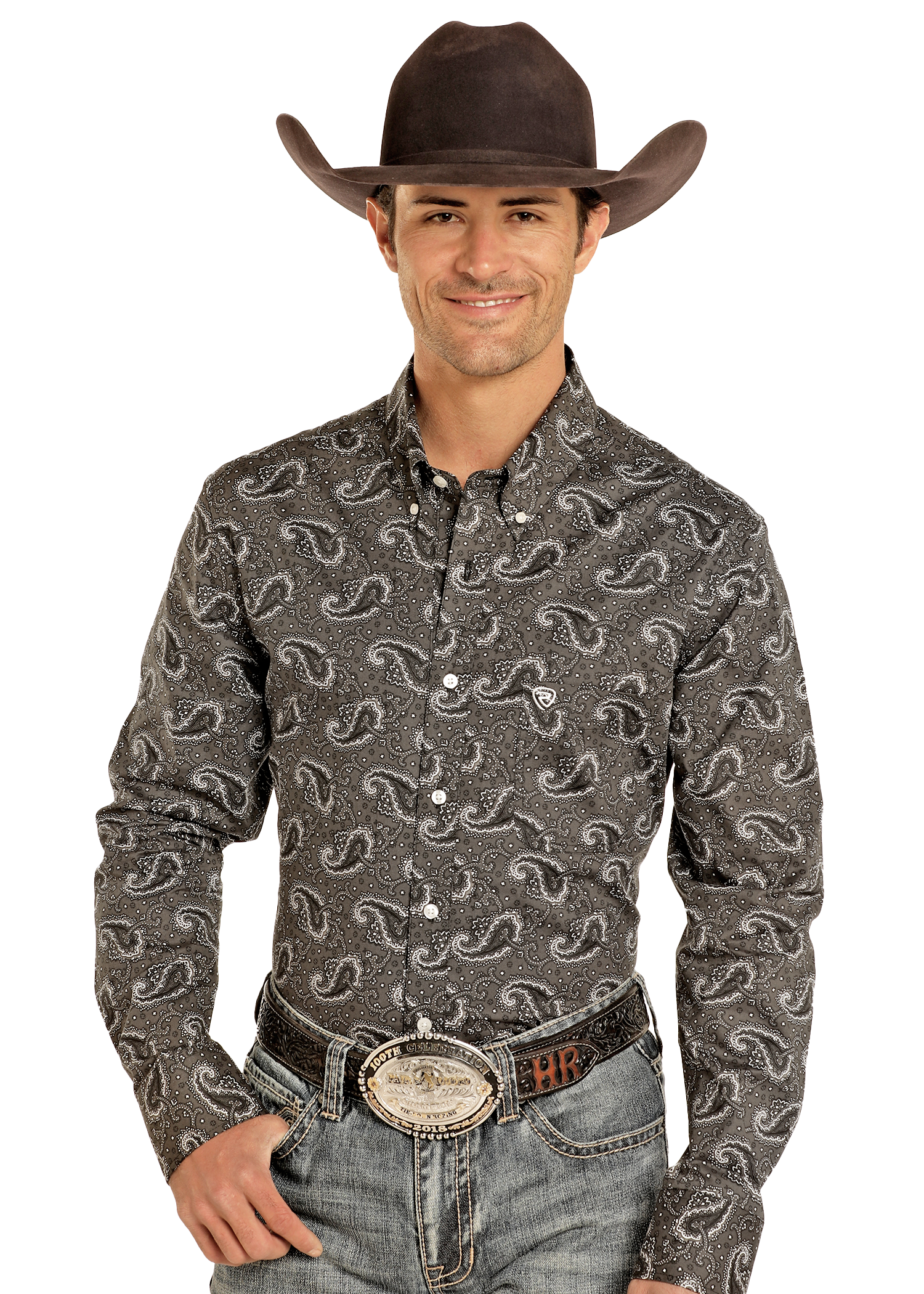 A man wearing a black cowboy hat, Rock & Roll Denim Classic Fit Paisley Print Long Sleeve Button Down Shirt, and blue jeans with a large belt buckle smiles with one hand in his pocket against a plain light background.