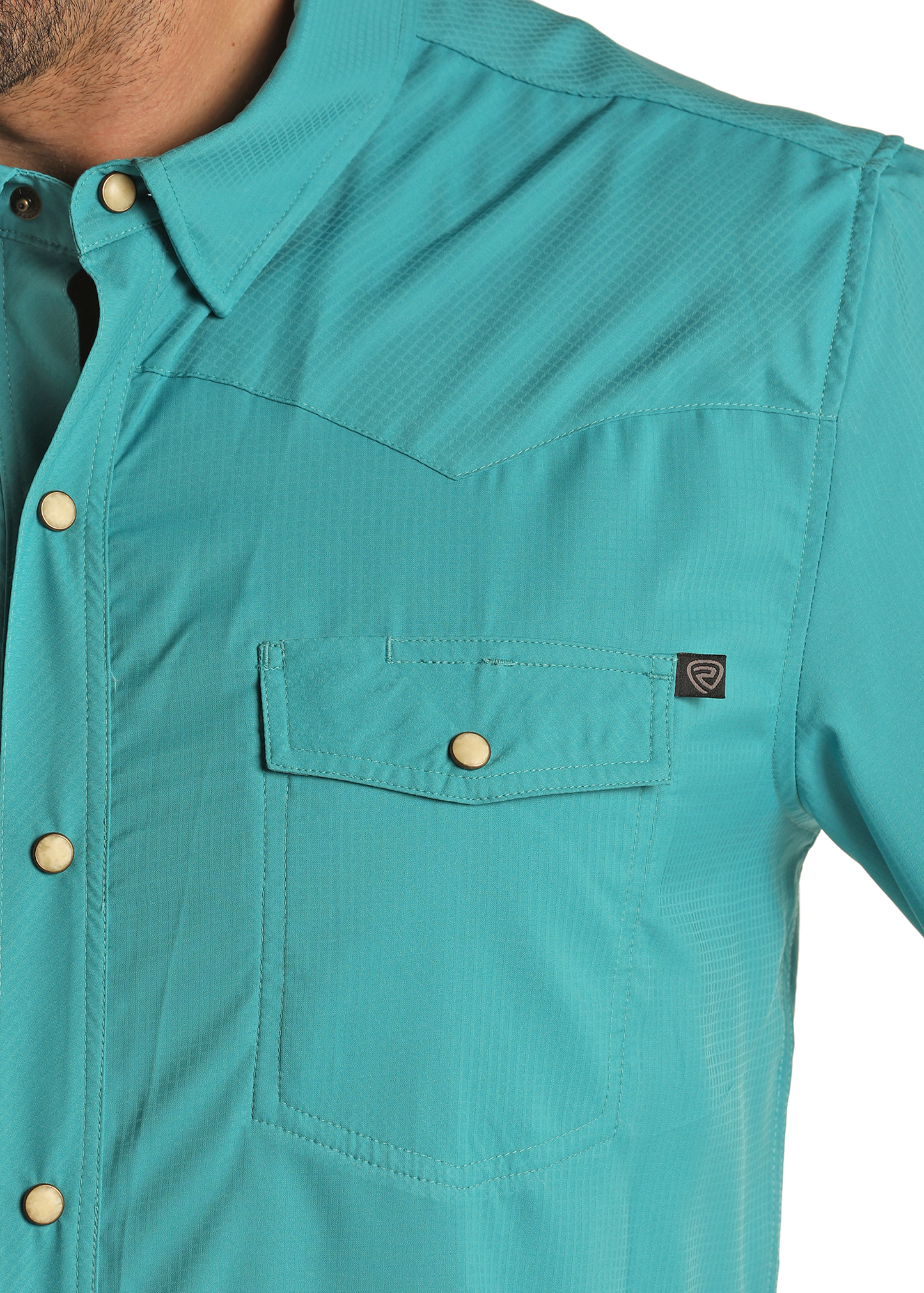 A close-up of the Rock & Roll Denim TEK Western Turquoise Long Sleeve Shirt shows gold snap buttons, a chest pocket with flap, and a small black tag. Part of the person's neck and shoulder are visible.