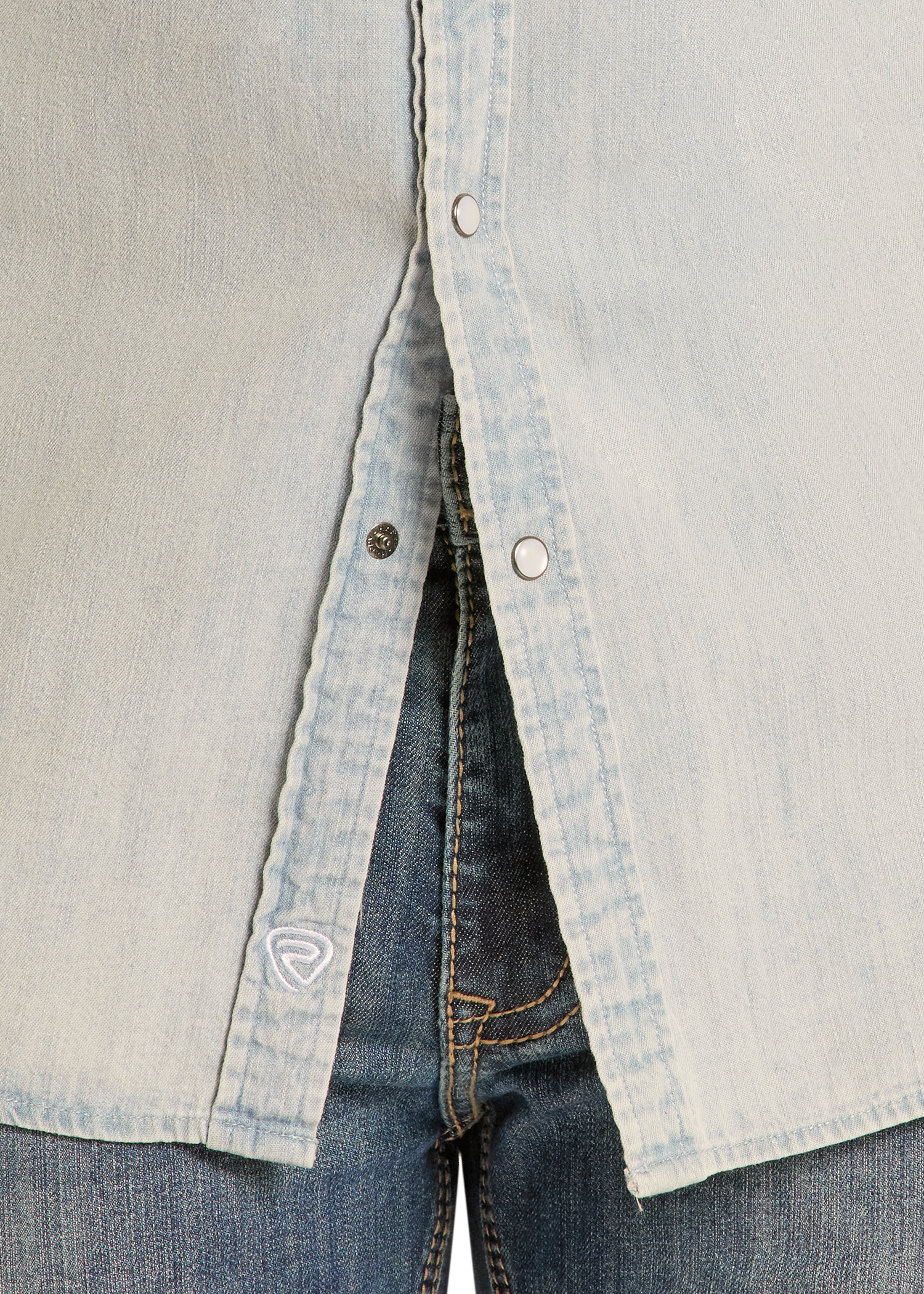 Close-up of the Rock & Roll Denim Slim Fit Light Wash Denim Snap Shirt, slightly open at the bottom to reveal blue jeans. Features snap buttons and a small white embroidered logo near the hem.