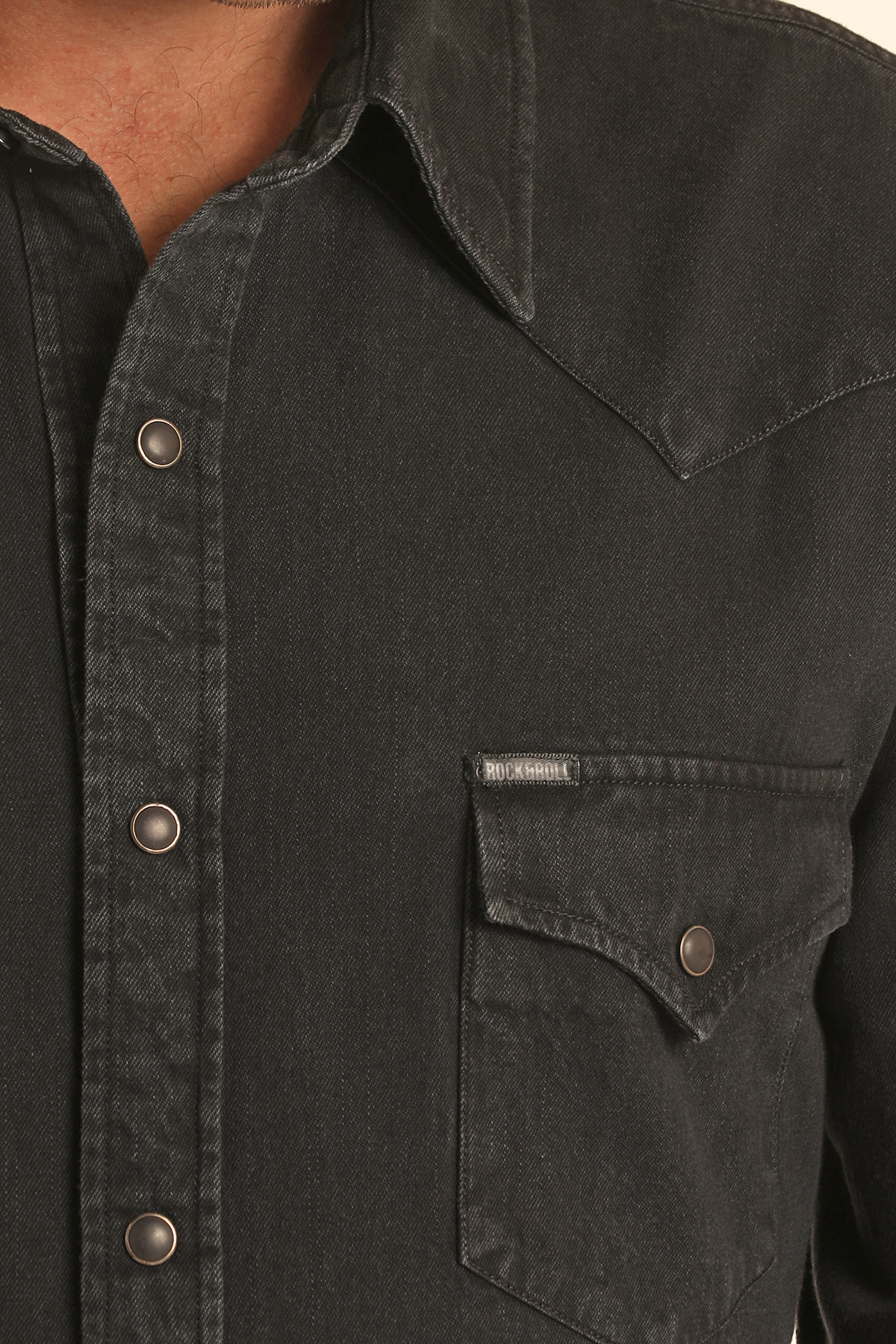 Close-up of the Rock & Roll Denim Slim Fit Black Wash Denim Snap Shirt, showing metal snap buttons, a flap chest pocket, prominent stitching, and a pointed collar. Only part of the wearer’s face and body are visible.
