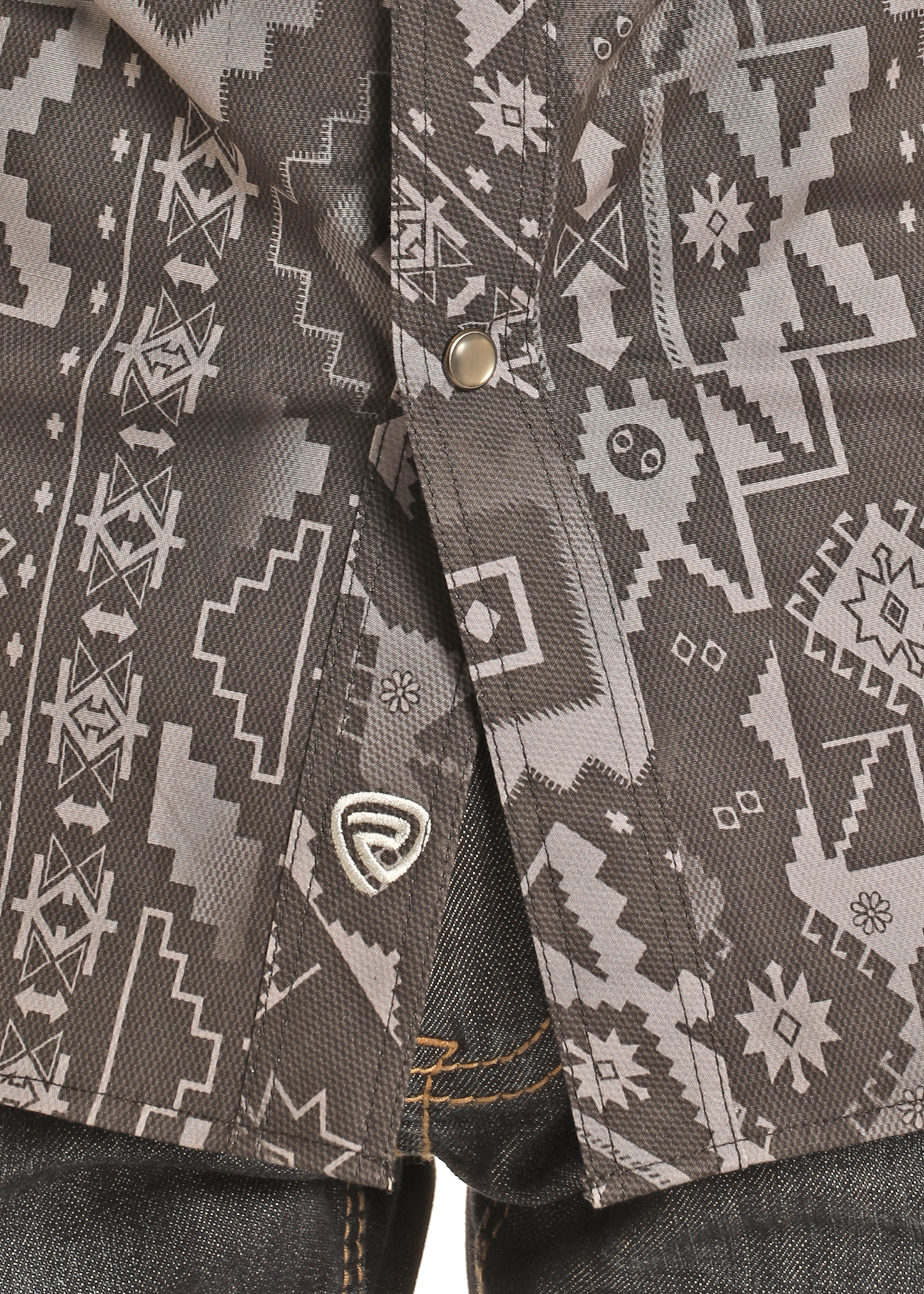 Close-up of a Rock & Roll Denim Slim Fit Black Aztec Print Long Sleeve Snap Shirt with white geometric and animal patterns, silver snaps, and metallic logo near the hem, partially covering dark jeans with orange stitching.