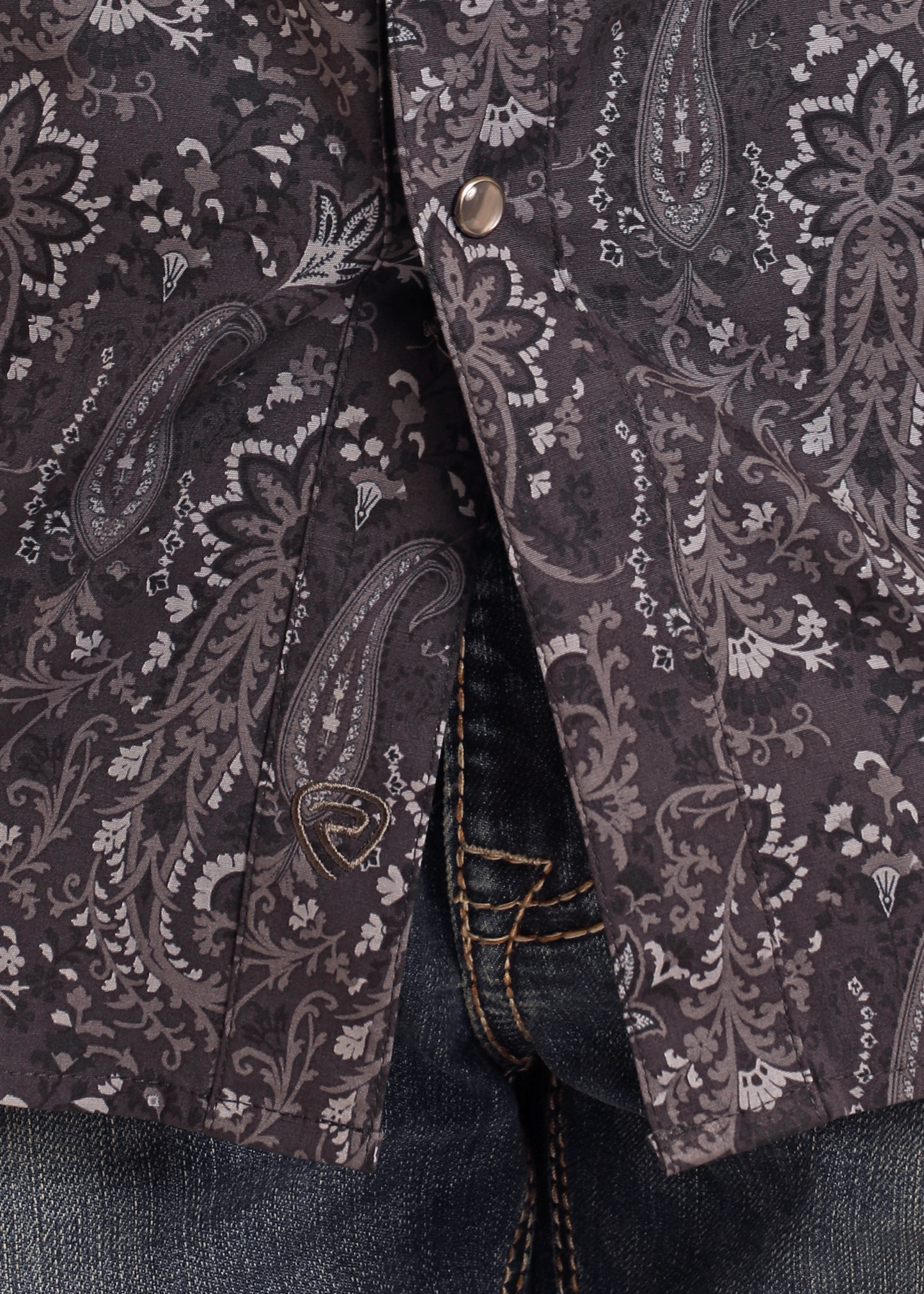 Close-up of the Rock & Roll Denim Slim Fit Black Paisley Print Long Sleeve Snap Shirt, featuring black and gray patterns with metal snap buttons, partially covering blue denim jeans with yellow stitching.