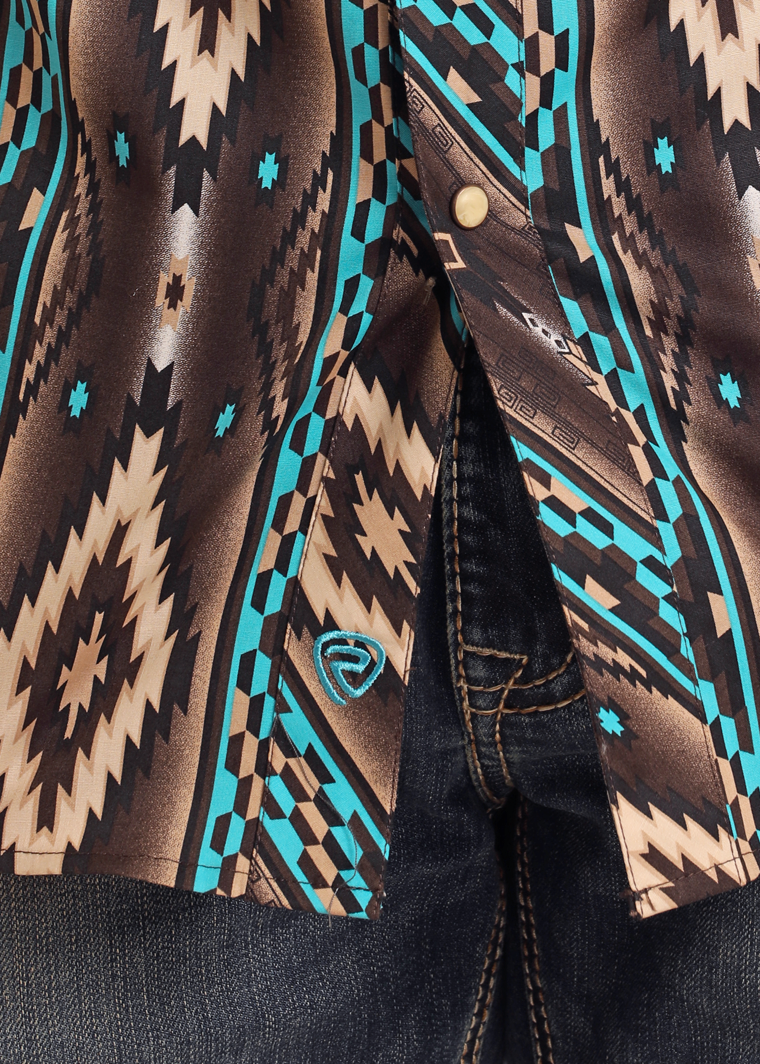 A close-up of the Rock & Roll Denim Slim Fit Southwestern Striped Long Sleeve Snap Shirt, featuring brown, beige, and turquoise geometric patterns tucked into dark blue jeans with a snap closure and a small blue embroidered logo near the hem.