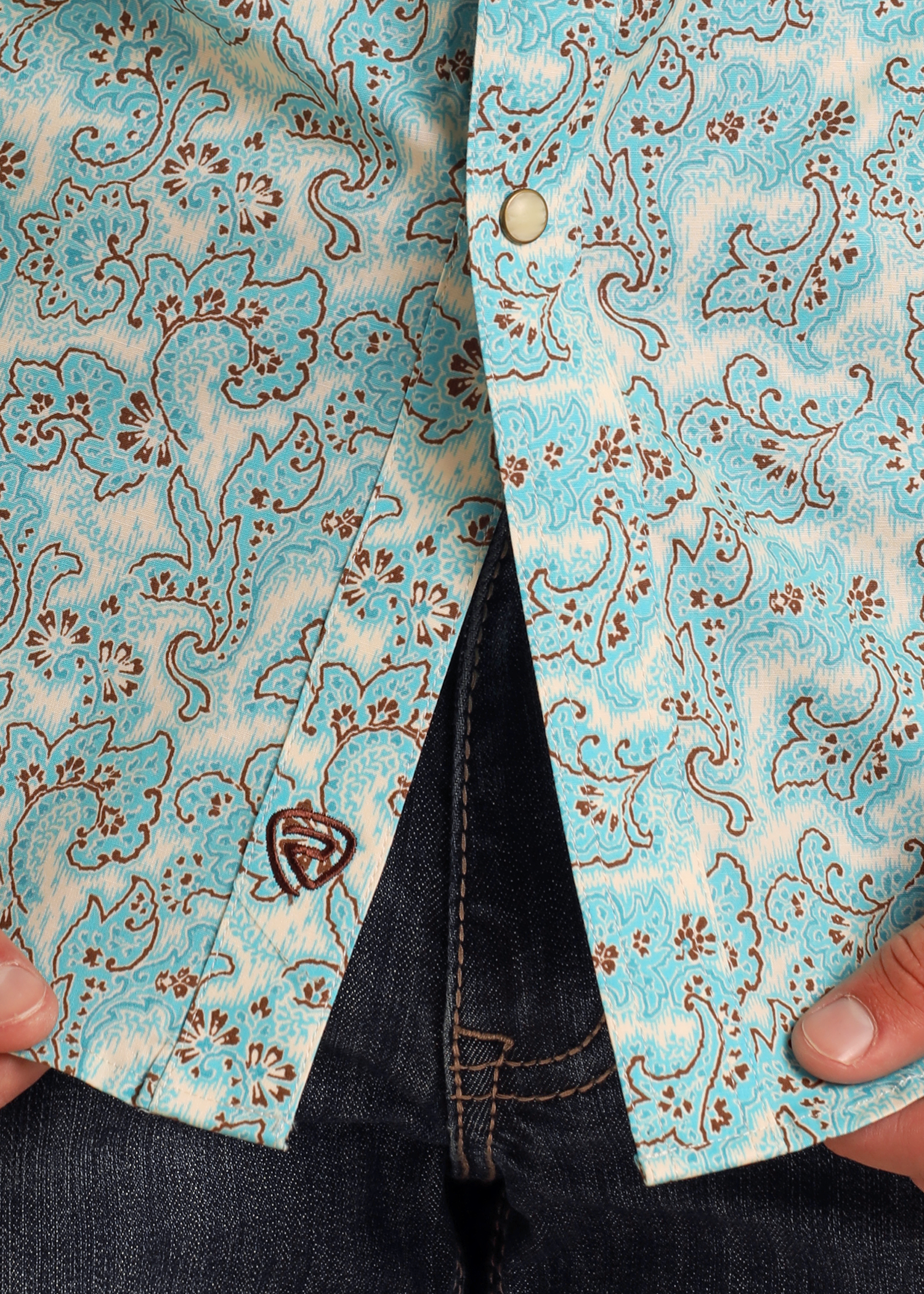 A person wears blue jeans and a Rock & Roll Denim Slim Fit Turquoise Paisley Print Long Sleeve Snap Shirt with brown floral designs. The shirt is unbuttoned at the bottom, partially revealing the jeans, with two hands near its edges.