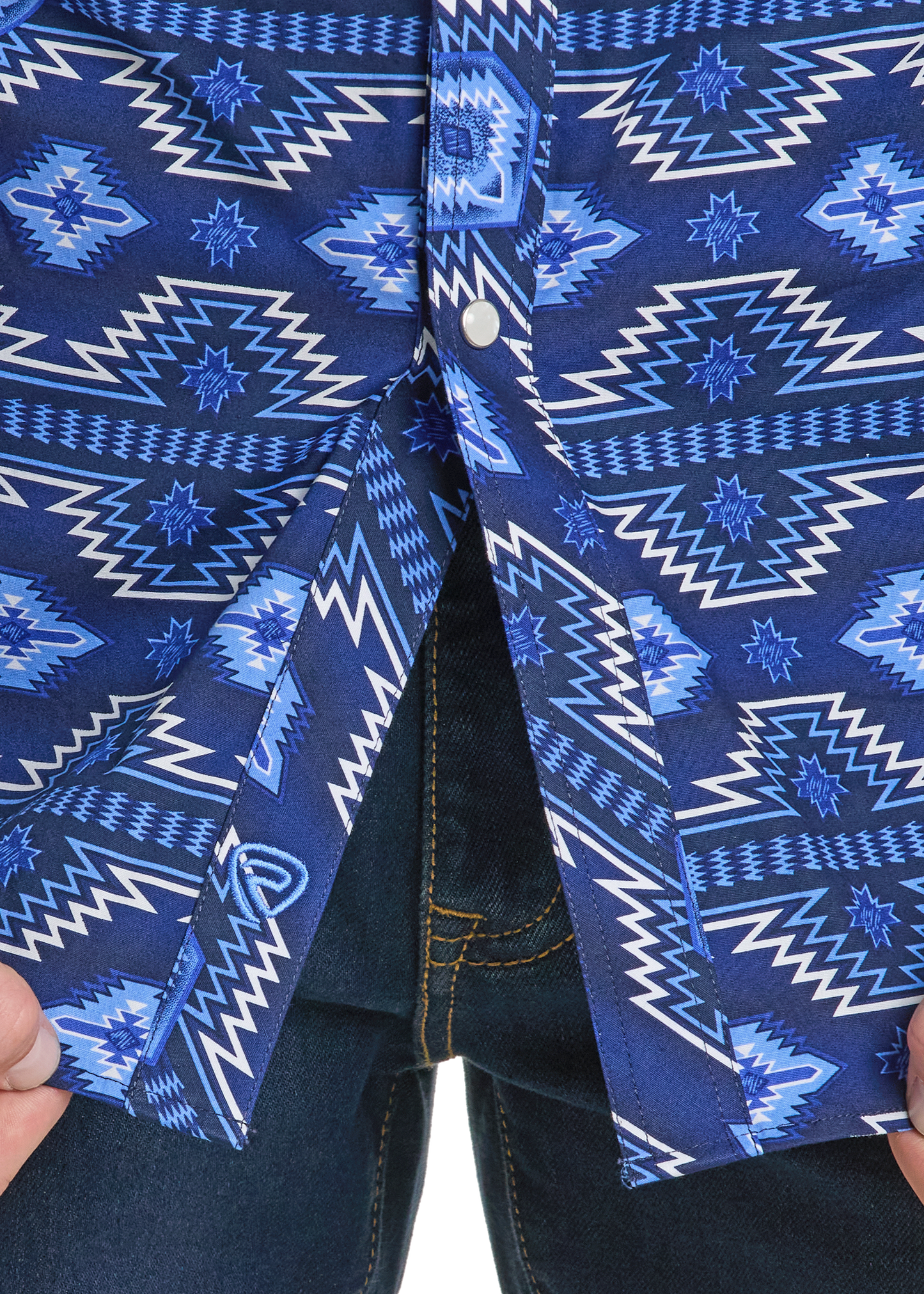 A close-up shows hands pulling apart the bottom of the Rock & Roll Denim Slim Fit Blue on Blue Aztec Long Sleeve Snap Shirt, revealing dark blue jeans. This men’s shirt features blue and white geometric patterns.
