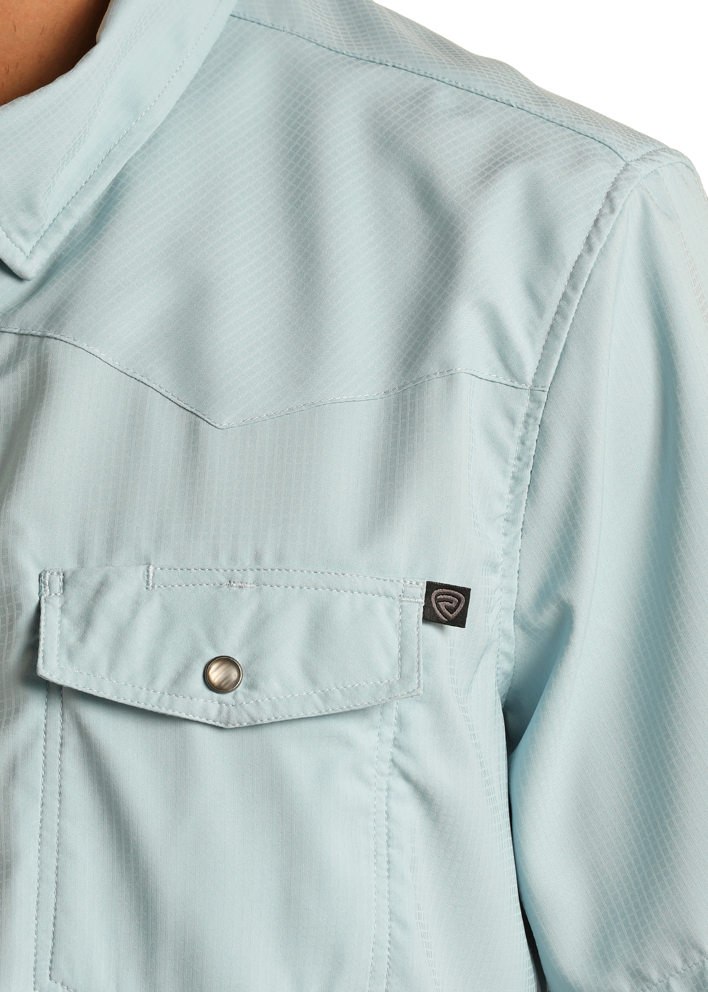 Close-up of the TEK Western Aqua Short Sleeve Shirt by Rock & Roll Denim, showing a buttoned chest pocket with a black tag. This light blue shirt features a subtle texture and offers moisture-wicking comfort.
