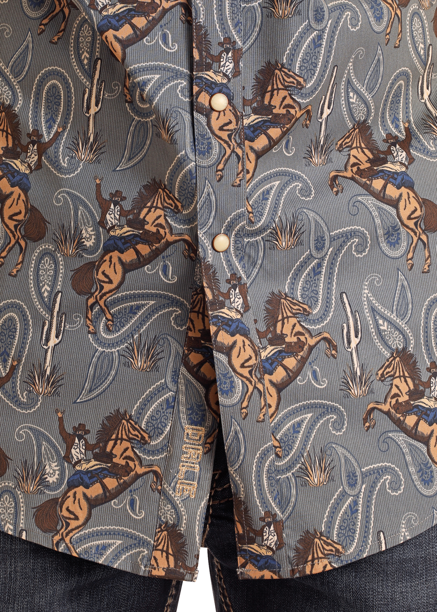 Closeup of the Rock & Roll Denim Dale Brisby Slim Fit Cowboy Print Short Sleeve Snap Shirt, featuring rodeo cowboys, bucking horses, cacti, paisley in blue, brown, and tan tones with pearl snaps; denim jeans are visible at the bottom.