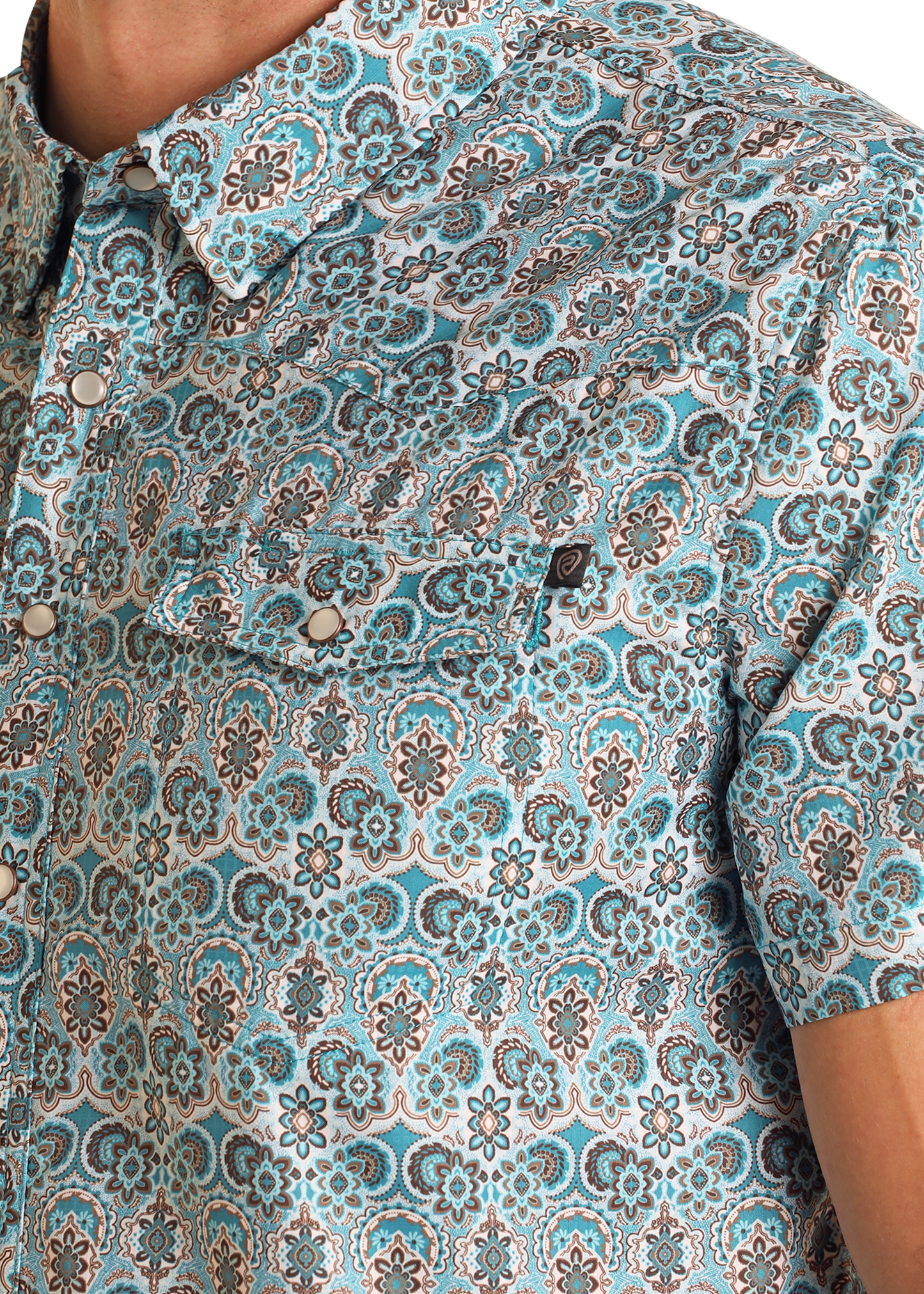 Close-up of a person wearing the TEK Western Geo Floral Print Short Sleeve Shirt by Rock & Roll Denim, showcasing blue, white, and brown intricate patterns. This short-sleeve button-up includes a front pocket with a button flap.