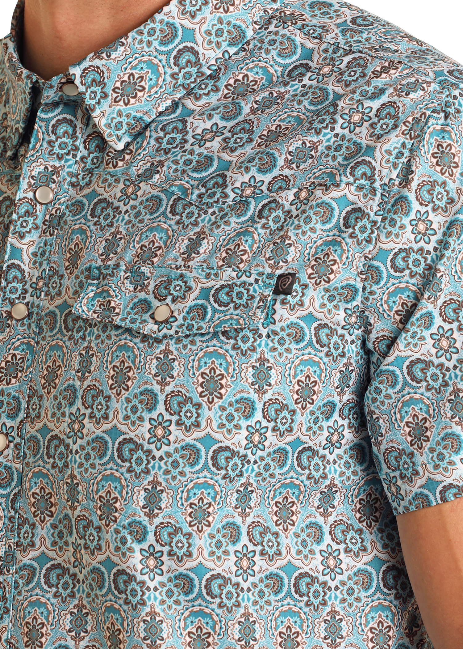 Close-up of a person wearing the TEK Western Geo Floral Print Short Sleeve Shirt by Rock & Roll Denim, showcasing blue, white, and brown intricate patterns. This short-sleeve button-up includes a front pocket with a button flap.