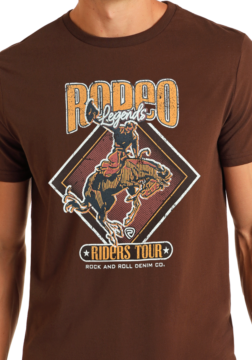 A person wears the Rodeo Legends Graphic Tee by Rock & Roll Denim, a comfortable brown shirt with a rodeo rider graphic and bold “RODEO Legends” and “RIDERS TOUR Rock & Roll Denim Co.” text.