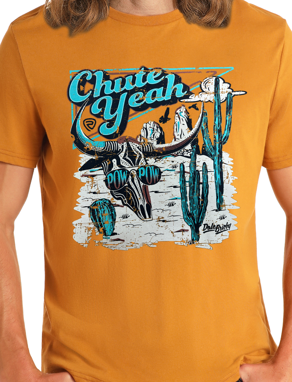 A person wearing the Rock & Roll Denim Dale Brisby Chute Yeah Graphic Tee in mustard yellow, featuring a bull skull, horns, cacti, desert scene, and blue and white “Chute Yeah” and “Pow Pow” text.
