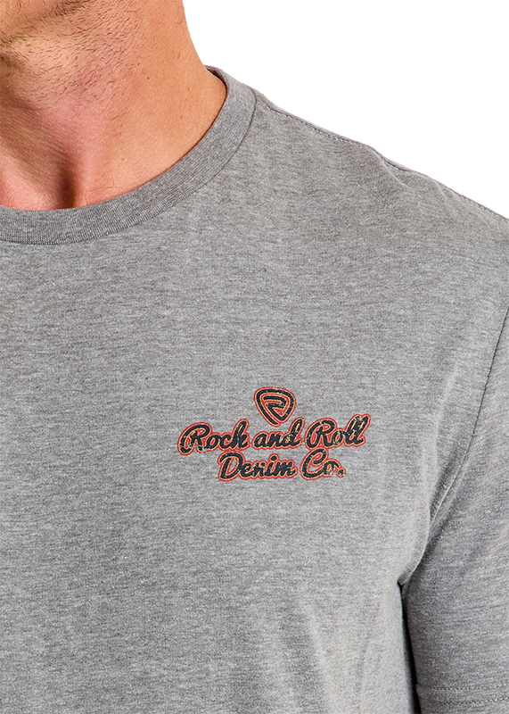 Close-up of a person wearing the Denim Co Unisex Graphic Tee by Rock & Roll Denim in light gray, featuring orange and brown embroidery on the left chest. The photo shows only the lower face, neck, and upper torso, highlighting its comfortable fit.