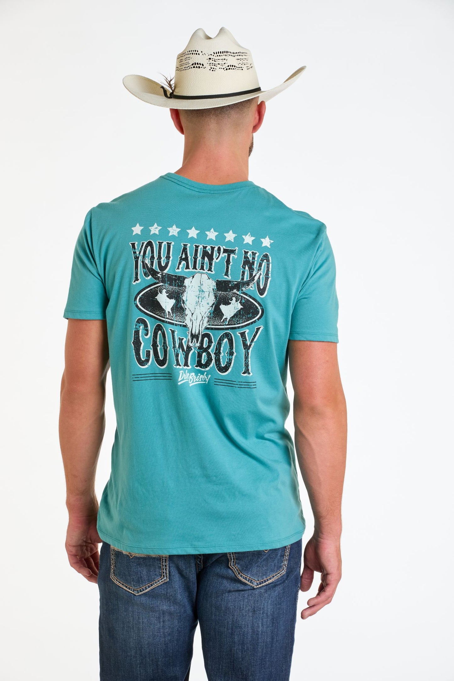 A man in a teal Rock & Roll Denim Dale Brisby You Ain't No Cowboy Graphic Tee, blue jeans, and cowboy hat stands facing away. The western shirt features YOU AIN'T NO COWBOY with a steer skull and stars on the back.