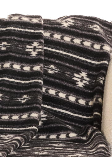 A black and off-white patterned knit throw blanket is draped over a cream-colored sofa, with a gray knit pillow on the left and a tall green cactus illustration on a white background.