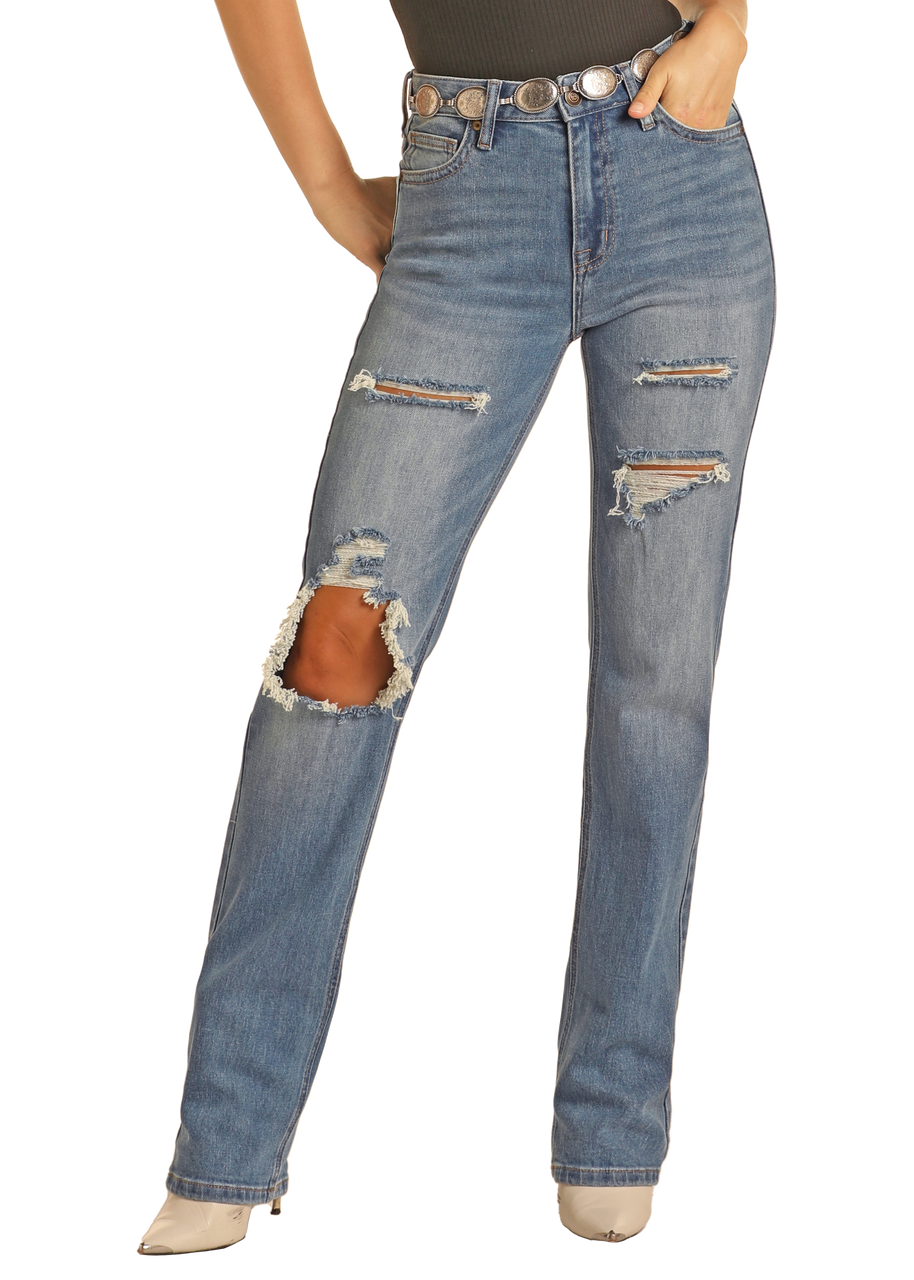 A person wears Rock & Roll Denim's West Desperado Distressed Denim High Rise Bootcut Jeans—a light blue cotton-poly blend with multiple rips, a large knee tear, paired with a silver-buckled belt and pointed-toe shoes.