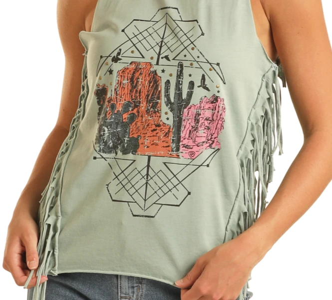 A person wearing the Rock & Roll Denim Desert Graphic Fringe Tank Top in light green, featuring western style, side fringe details, and a desert-themed print with cacti, rock formations, and geometric accents.