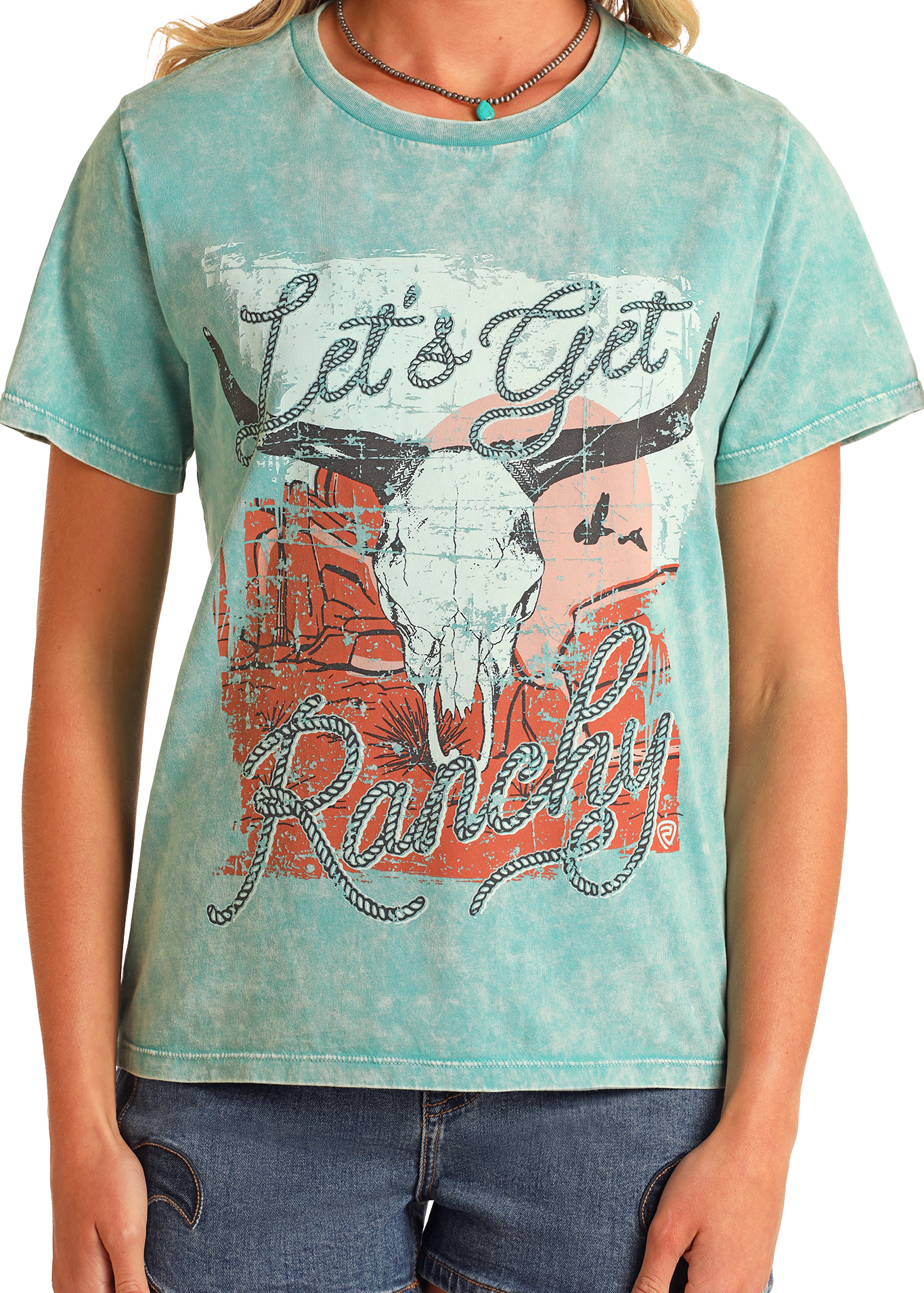 A person wearing the Rock & Roll Denim Let's Get Ranchy Graphic Tee in turquoise mineral-wash cotton, styled with denim shorts and a turquoise necklace for a Western vibe. Featuring a cow skull, desert scene, and lasso-style font.