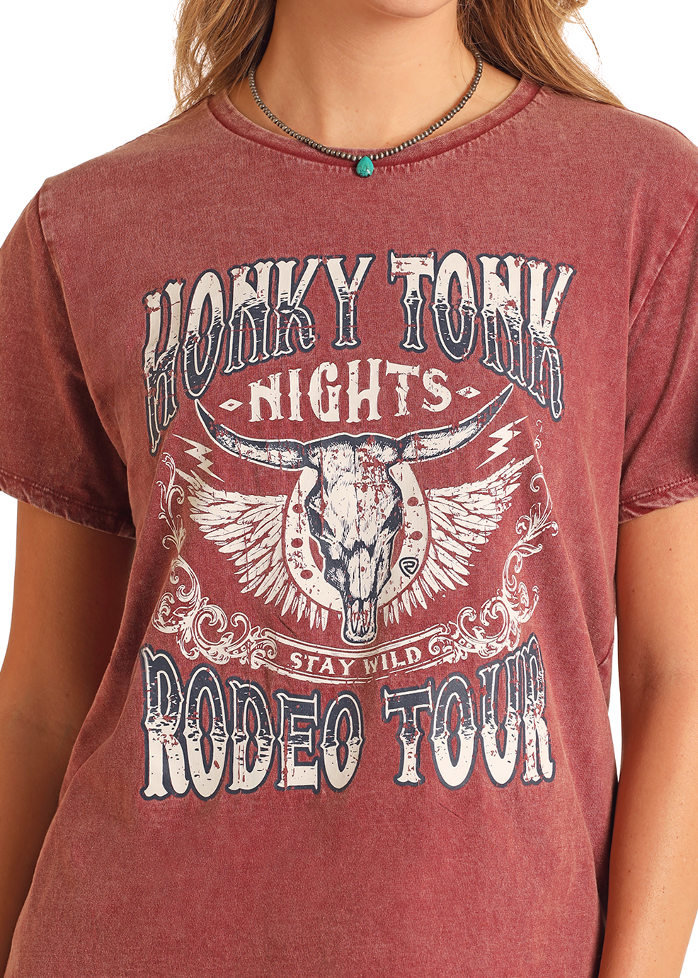 A person wears the Rock & Roll Denim Honky Tonk Nights Graphic Tee, featuring a western steer skull with wings design in faded red, paired with a necklace featuring a turquoise bead.