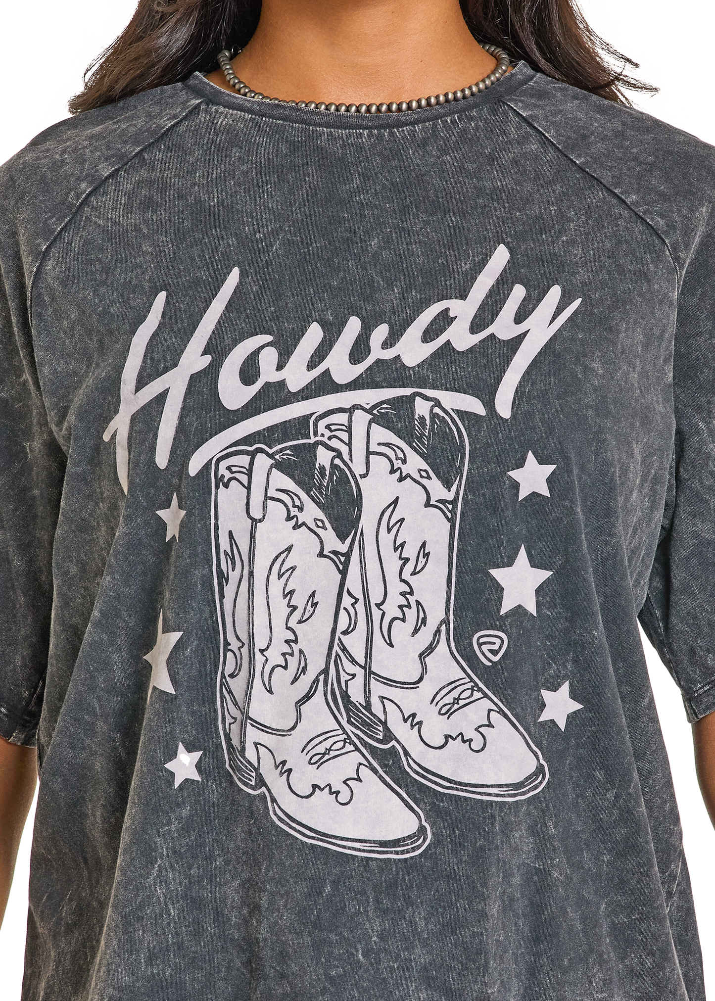 Wearing the Rock & Roll Denim Acid Wash Howdy Oversized Tee, which has "Howdy" above illustrated cowboy boots and white stars. Photo shows only the torso and part of a necklace in this boot graphic tee look.