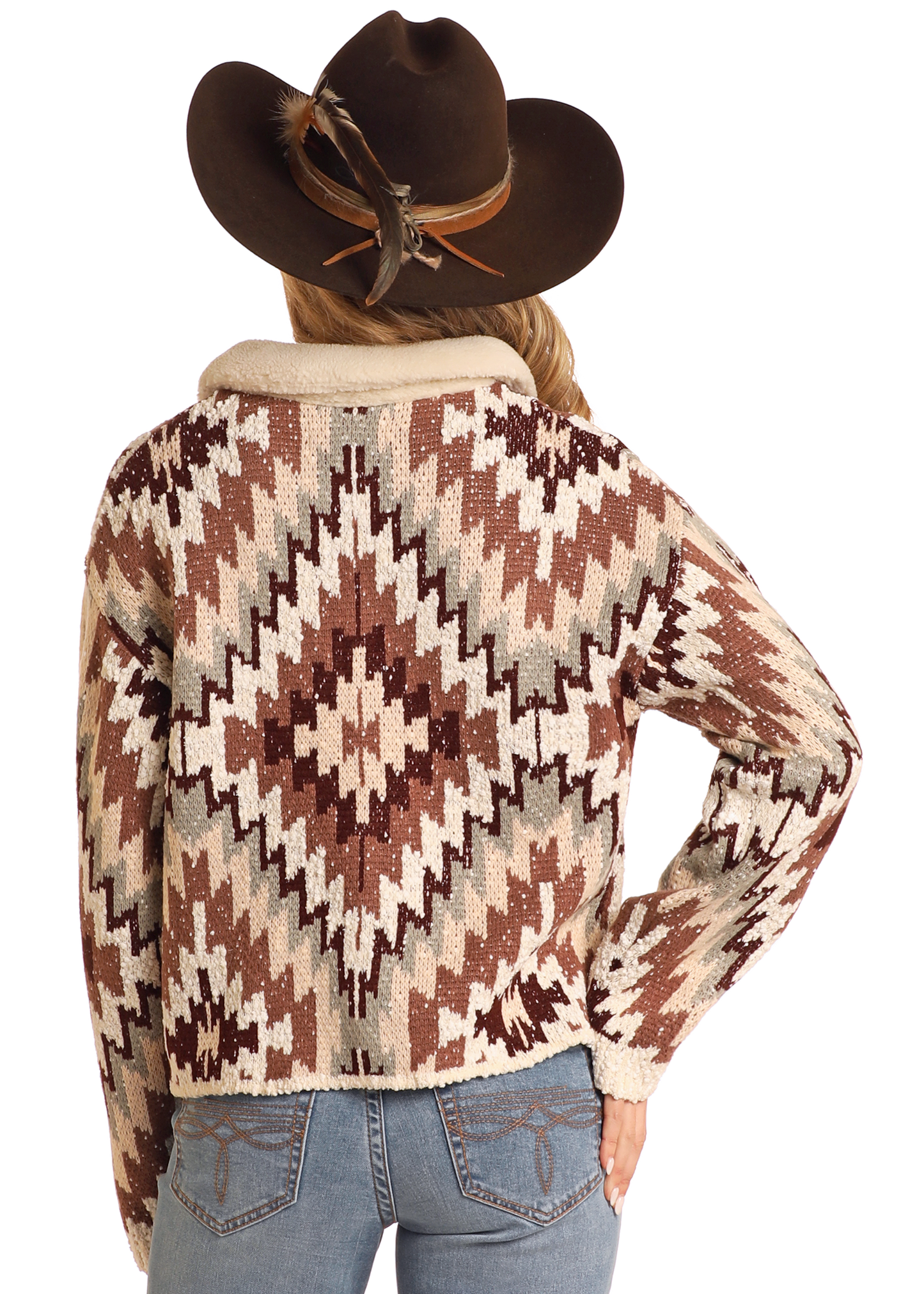 A person wearing a brown cowboy hat with a feather and an Aztec Button Up Sweater Cardigan by Rock & Roll Denim stands with their back to the camera, hand on hip, paired with light blue jeans.