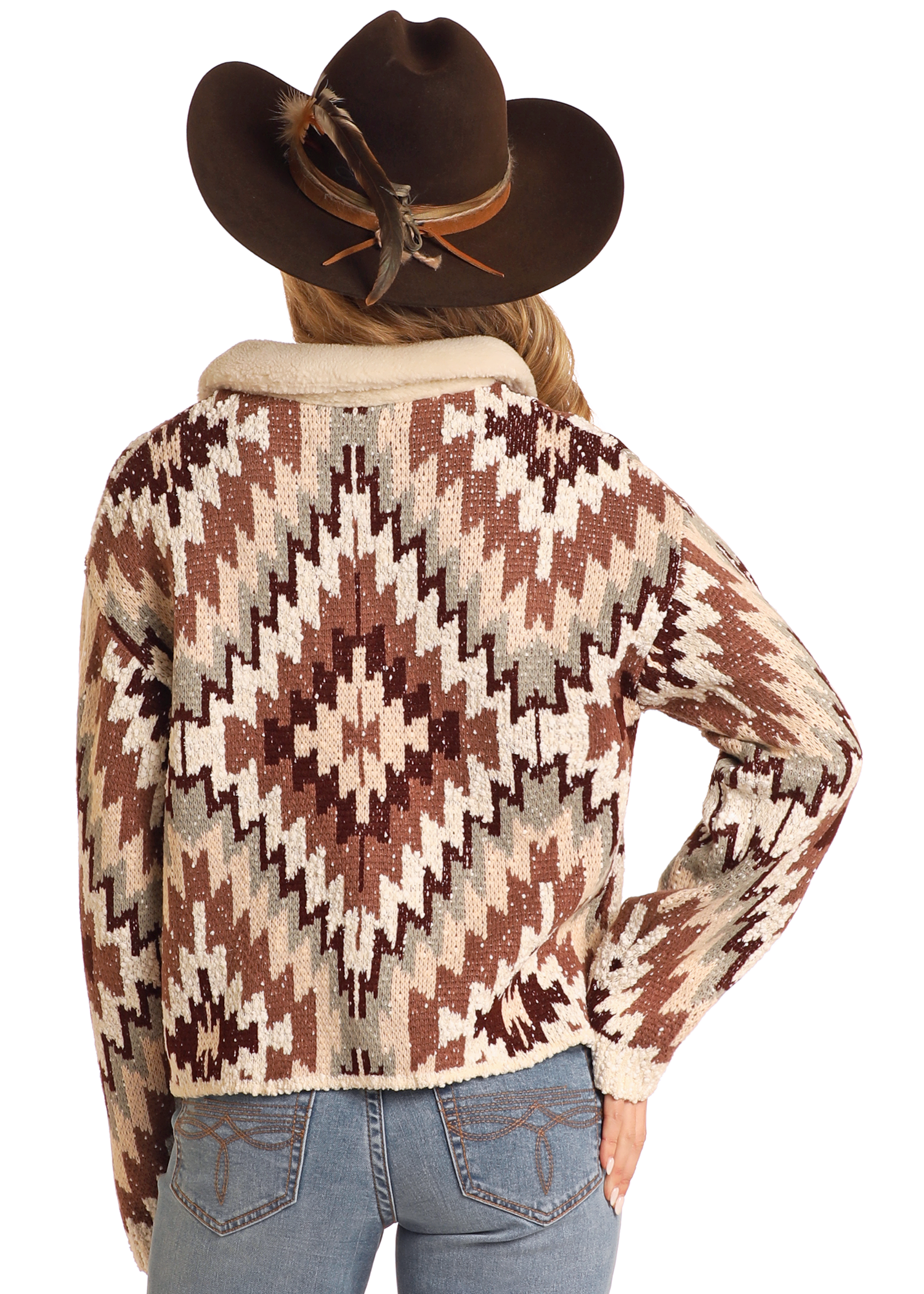 A person wearing a brown cowboy hat with a feather and an Aztec Button Up Sweater Cardigan by Rock & Roll Denim stands with their back to the camera, hand on hip, paired with light blue jeans.