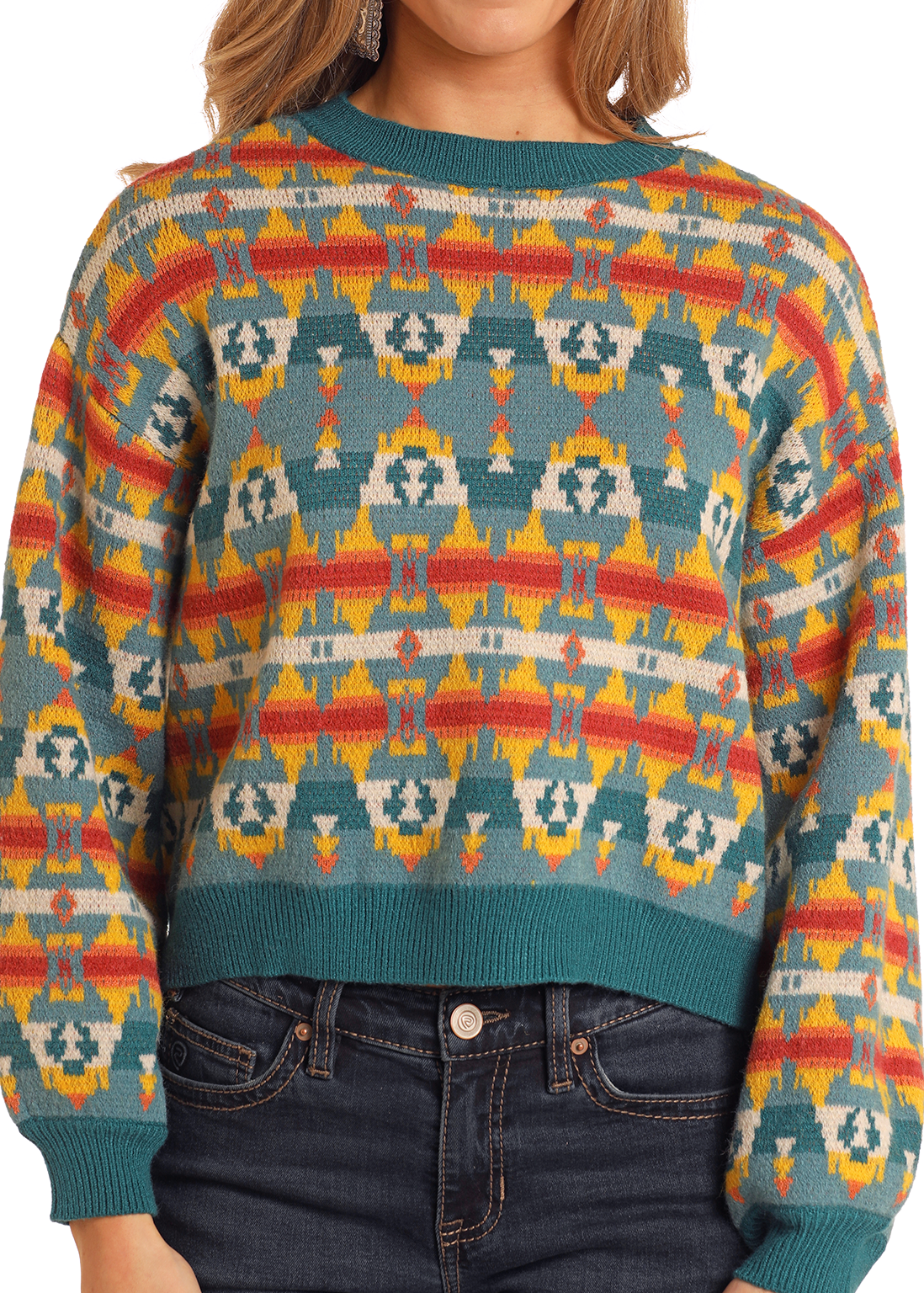 A woman models the Rock & Roll Denim Turquoise Aztec Cropped Sweater featuring vibrant teal, yellow, orange, and red geometric patterns. Paired with dark blue jeans, the photo shows her from shoulders to hips.