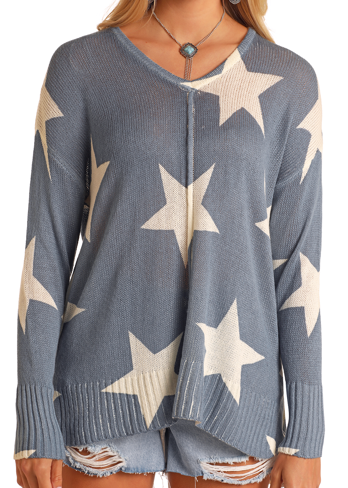 A woman wears the Rock & Roll Denim Star Print Sweater in blue with large white stars, made from soft acrylic, styled with distressed denim shorts and a turquoise stone necklace.