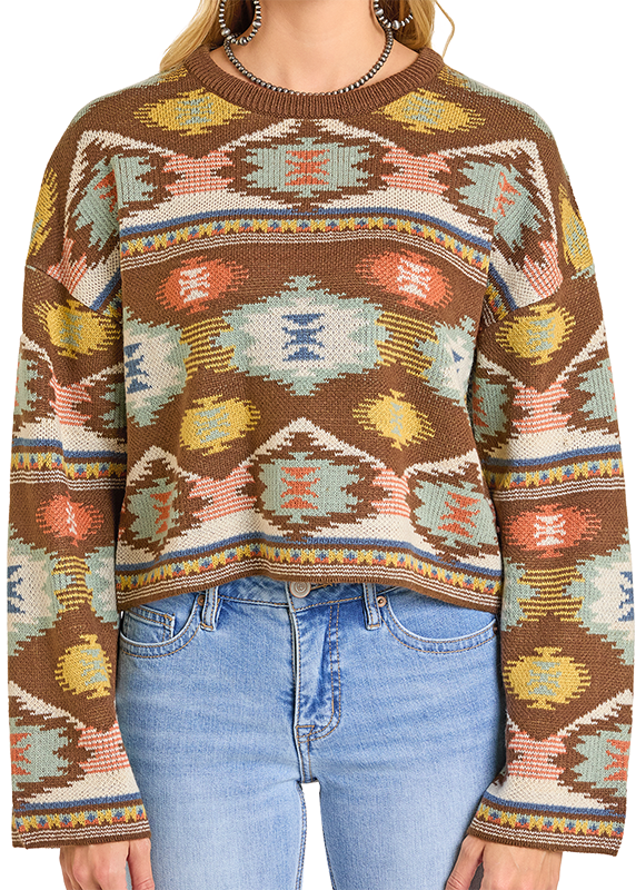 A person wearing a Rock & Roll Denim Aztec Cropped Sweater with a colorful teal, yellow, and white geometric pattern, long sleeves, and a relaxed cropped fit, paired with light blue jeans.