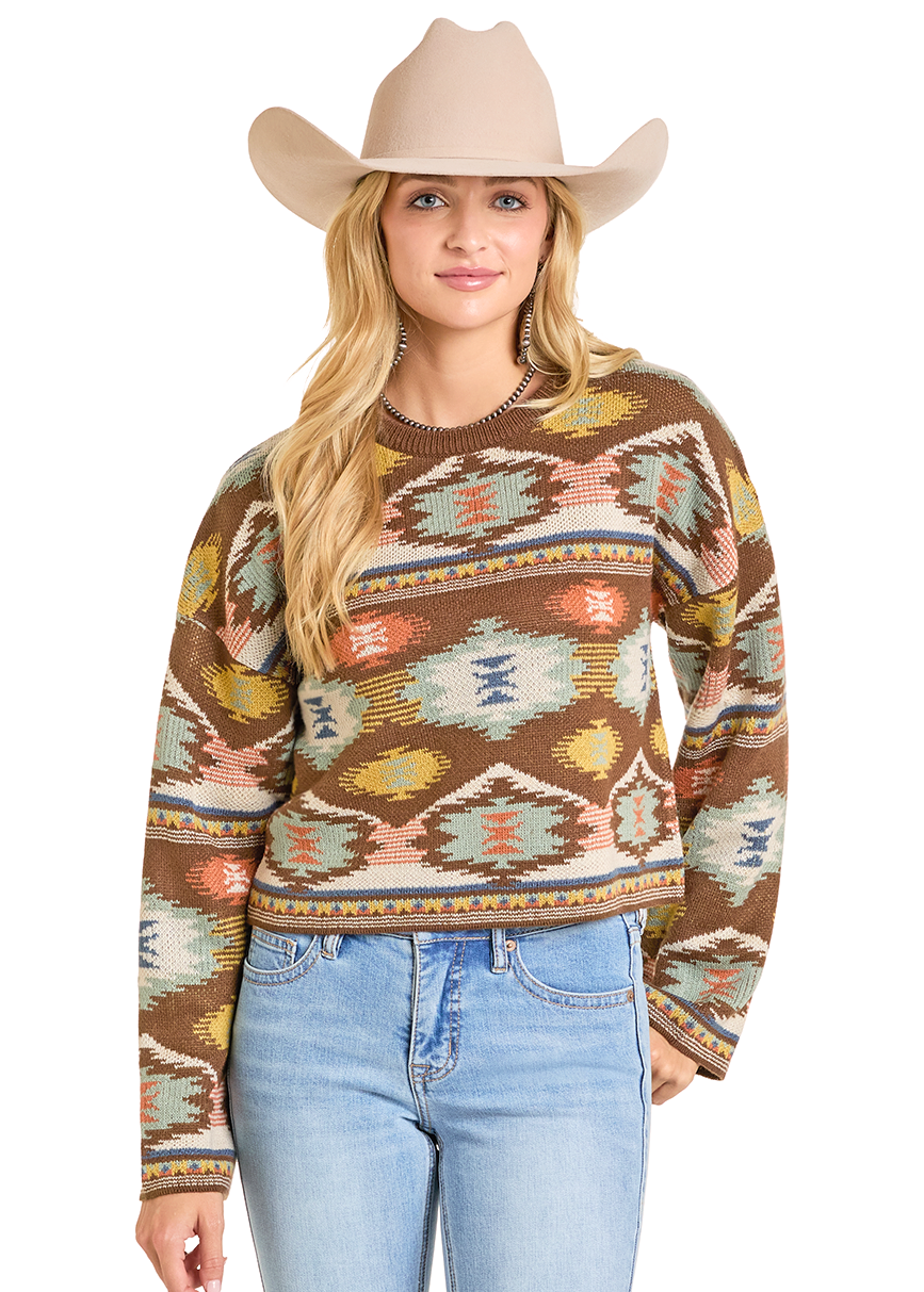 A woman in a beige cowboy hat, light blue jeans, and the Rock & Roll Denim Aztec Cropped Sweater with colorful geometric designs stands confidently, smiling with one hand in her pocket against a plain white background.